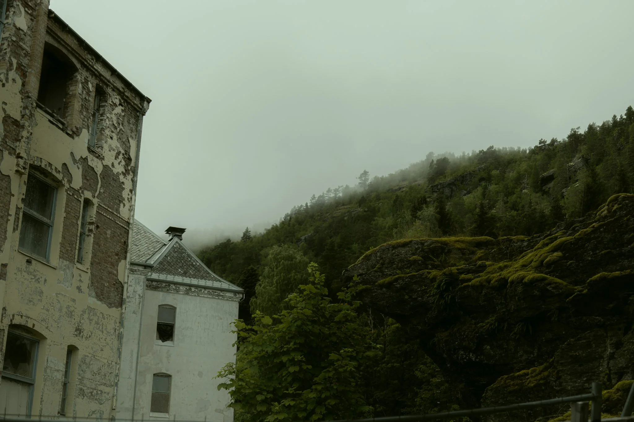 An old, weathered building with peeling paint and damaged walls next to a lush, green hillside shrouded in fog or low clouds.