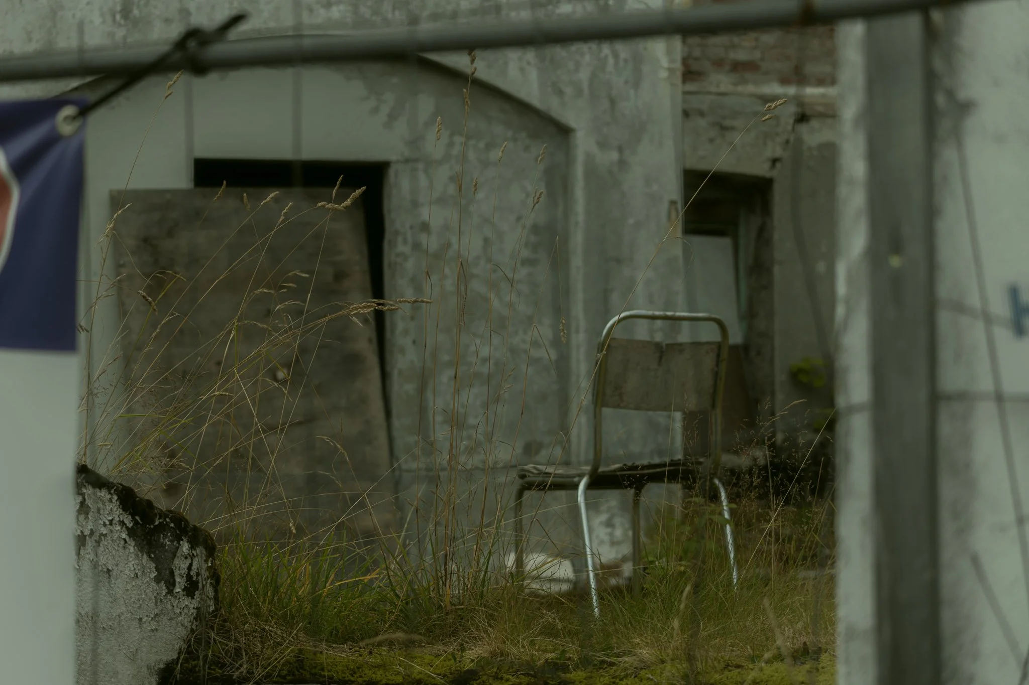 Overgrown yard with dry grass, a cement block wall, and a folding chair in front of a concrete building with unfinished surfaces.