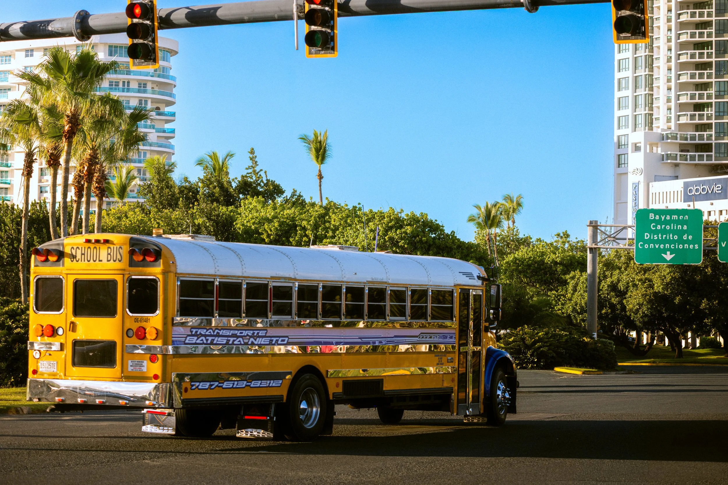 A yellow school bus in front of tall modern buildings with balconies, trees, and road signs in a sunny urban setting.