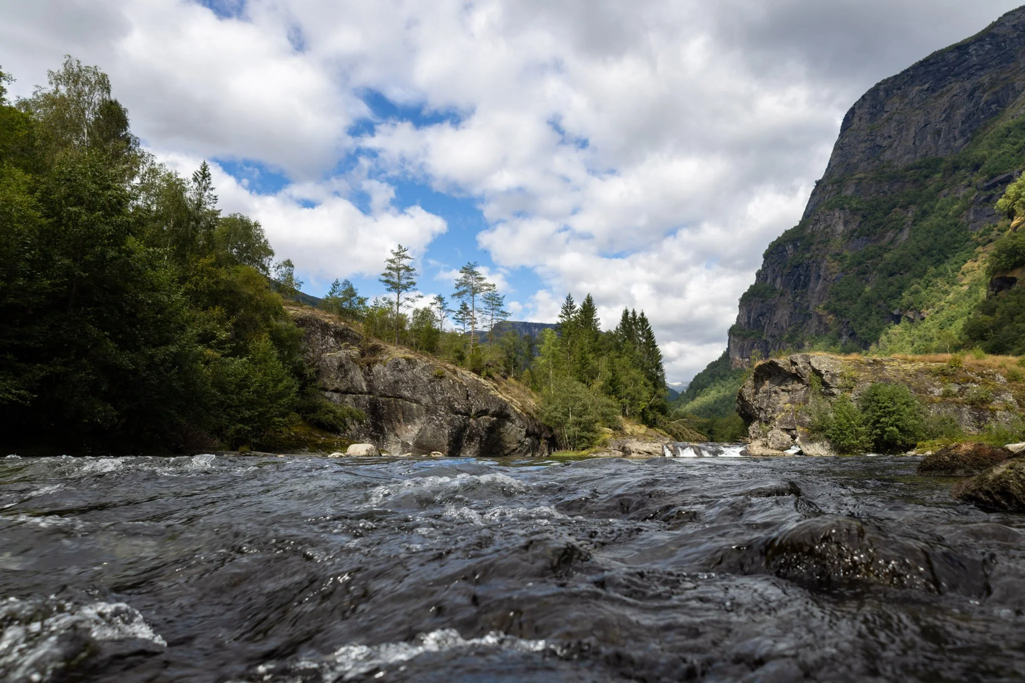 A wide river flowing through a mountainous landscape with a cloudy sky overhead, surrounded by lush green trees and rocky cliffs.