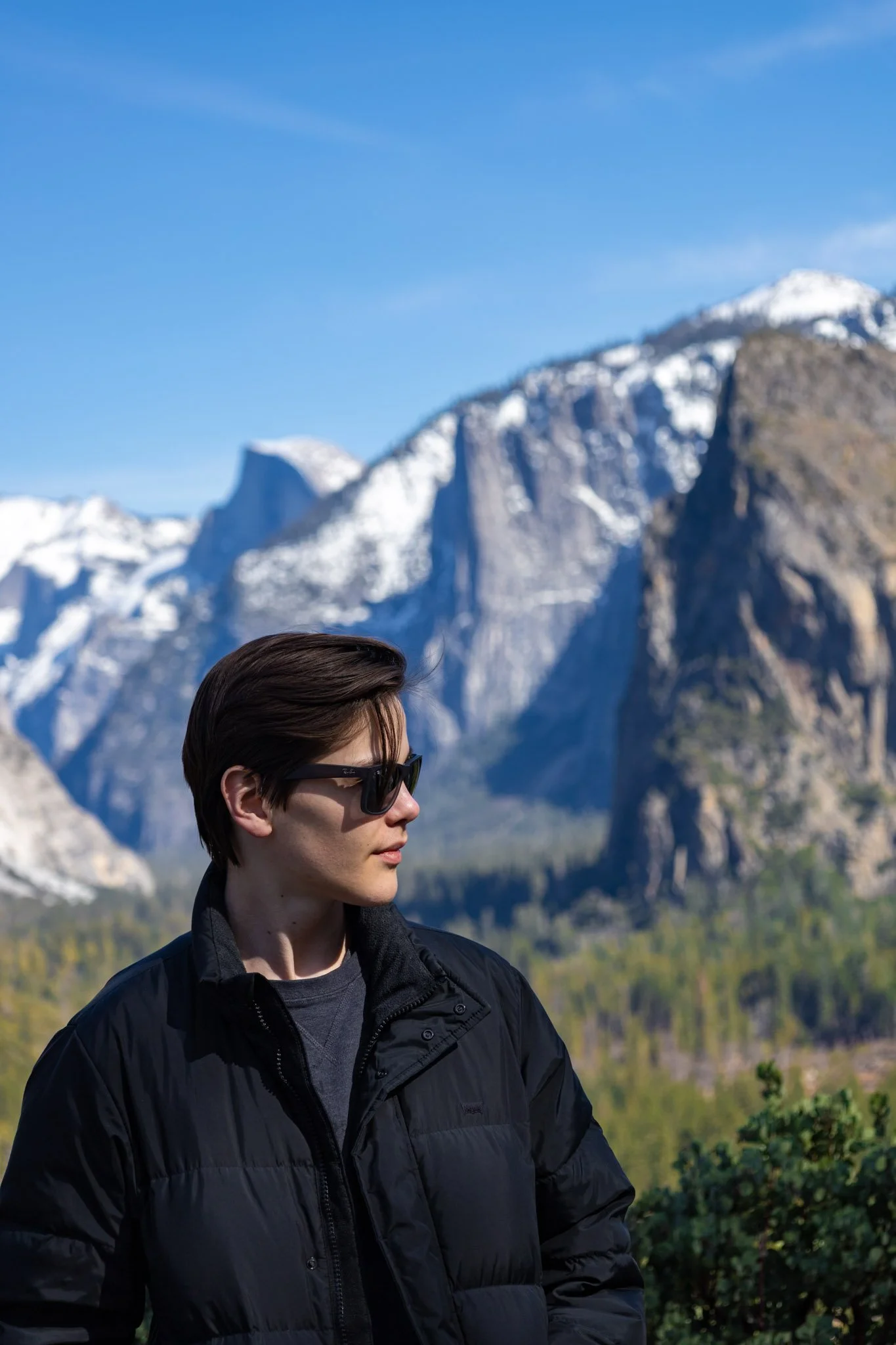 A young man with dark hair wearing sunglasses and a black jacket standing outdoors with mountains and a clear blue sky in the background.
