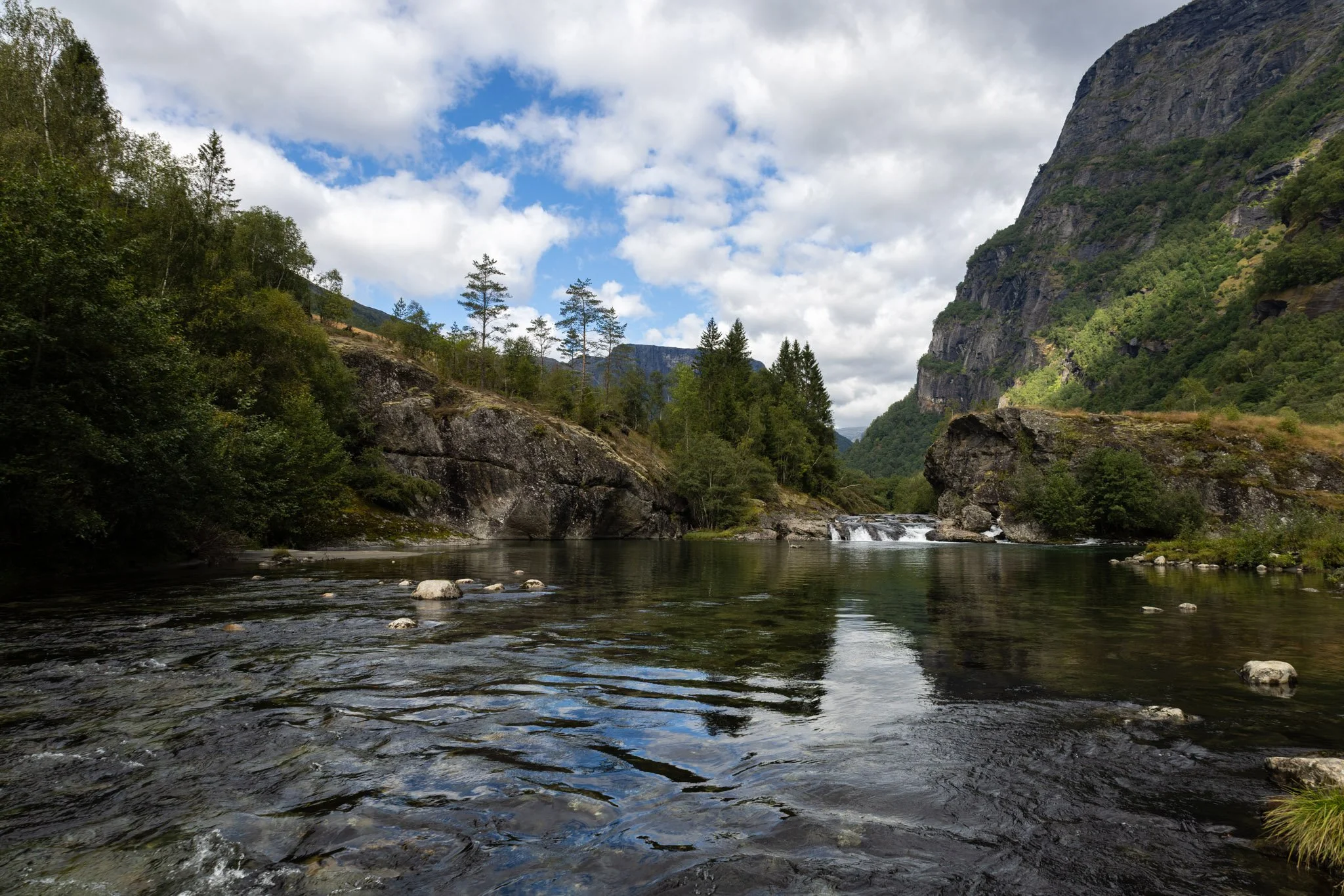 A river flowing through a mountainous landscape with trees, rocky cliffs, and a partly cloudy sky.