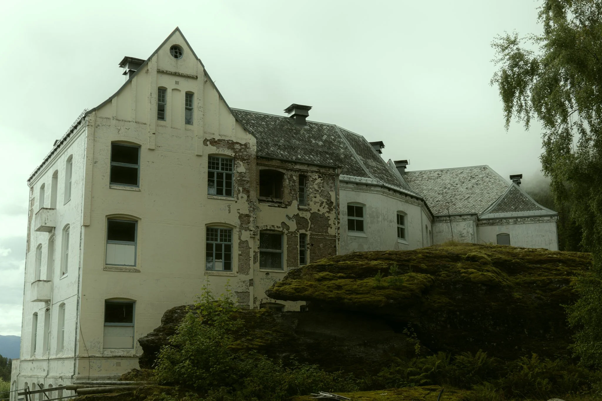 An old, weathered white house with peeling paint and damaged sections, surrounded by greenery and moss-covered rocks, under a cloudy sky.