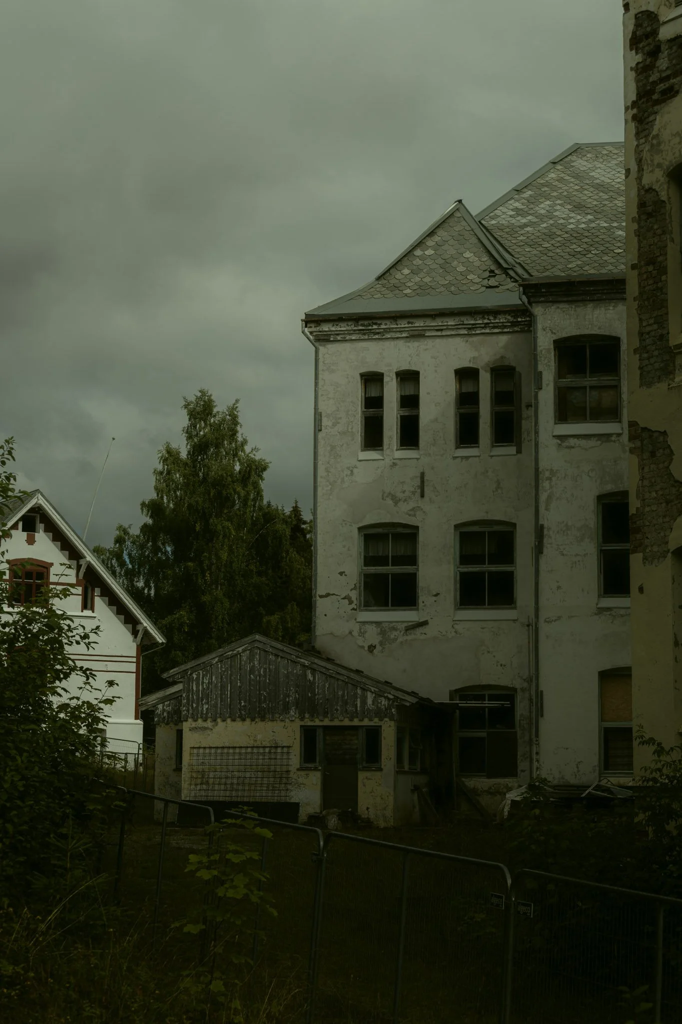 Old multi-story building with weathered white walls and multiple windows, next to a smaller wooden structure, under a cloudy sky with surrounding trees.