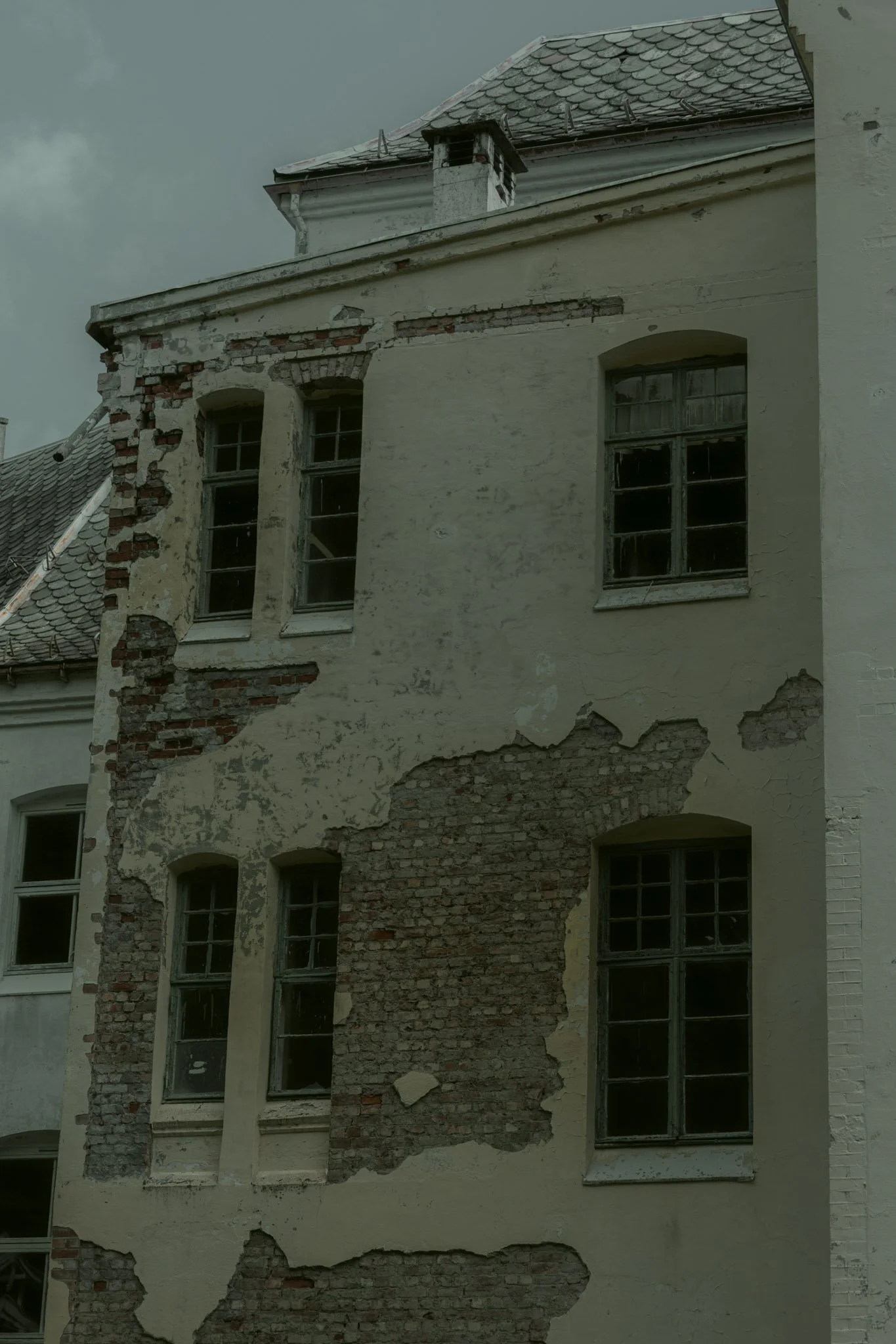 A building with partially damaged walls and peeling paint, revealing bricks underneath. The building has tall, narrow windows with some broken or missing glass, and a tiled roof.