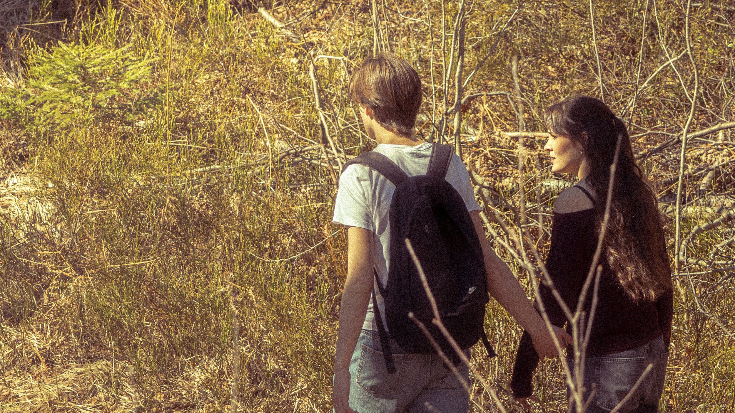 A young man and woman walking through a natural, dry, brushy area during daytime, holding hands, both with backpacks.