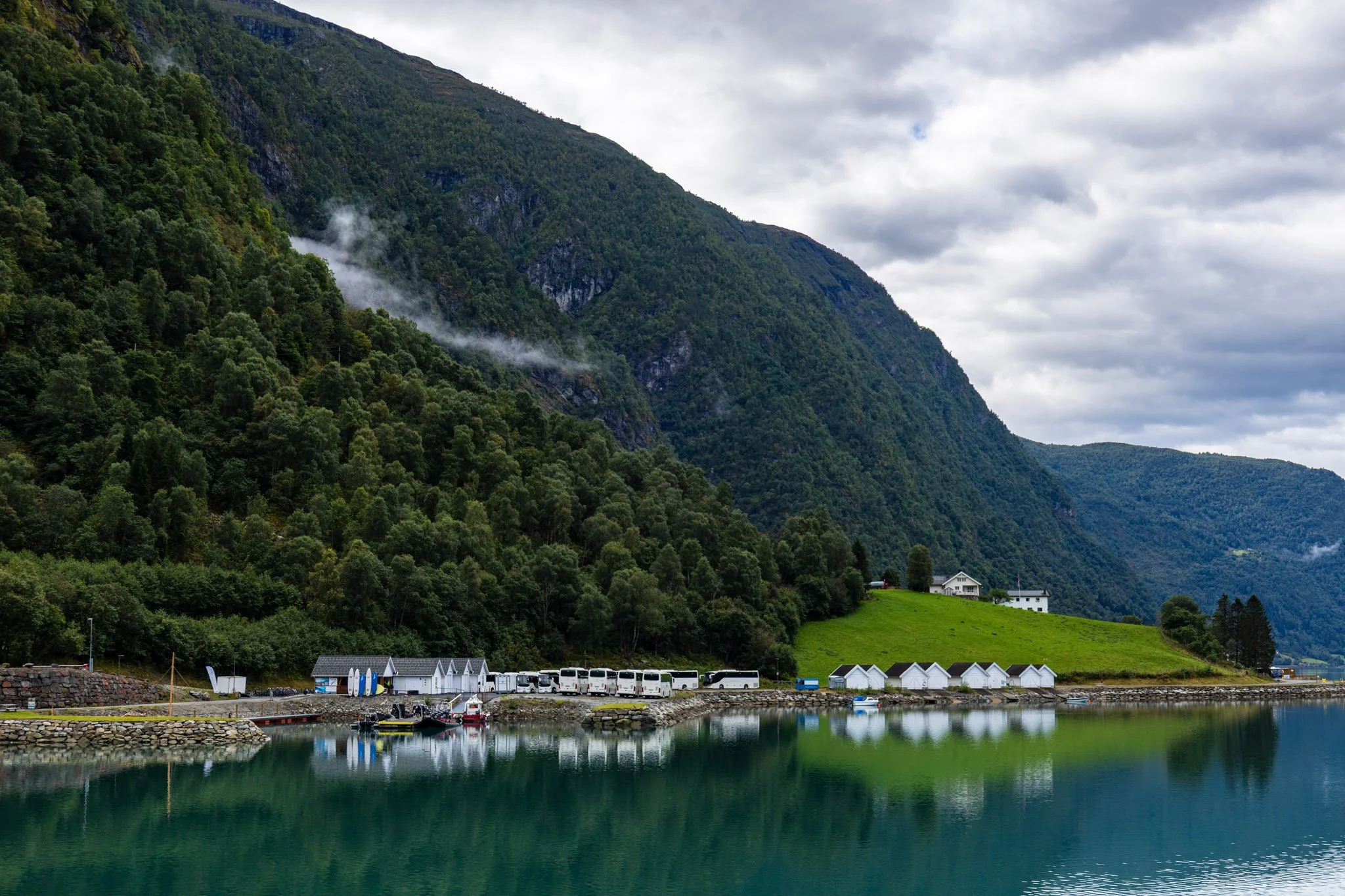 Scenic view of a fjord with calm water reflecting the cloudy sky, a row of white boats along the shoreline, green grassy hills, and houses with mountains covered in dense forest in the background.