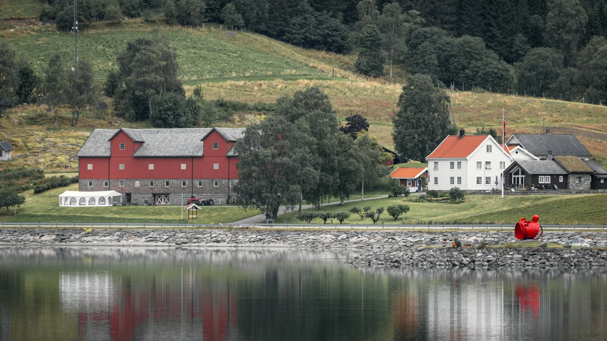 Scenic view of a lakeside area featuring houses, including a red barn and white house, with greenery and trees on surrounding hills in the background. There is a small white tent and a red sculpture near the water.