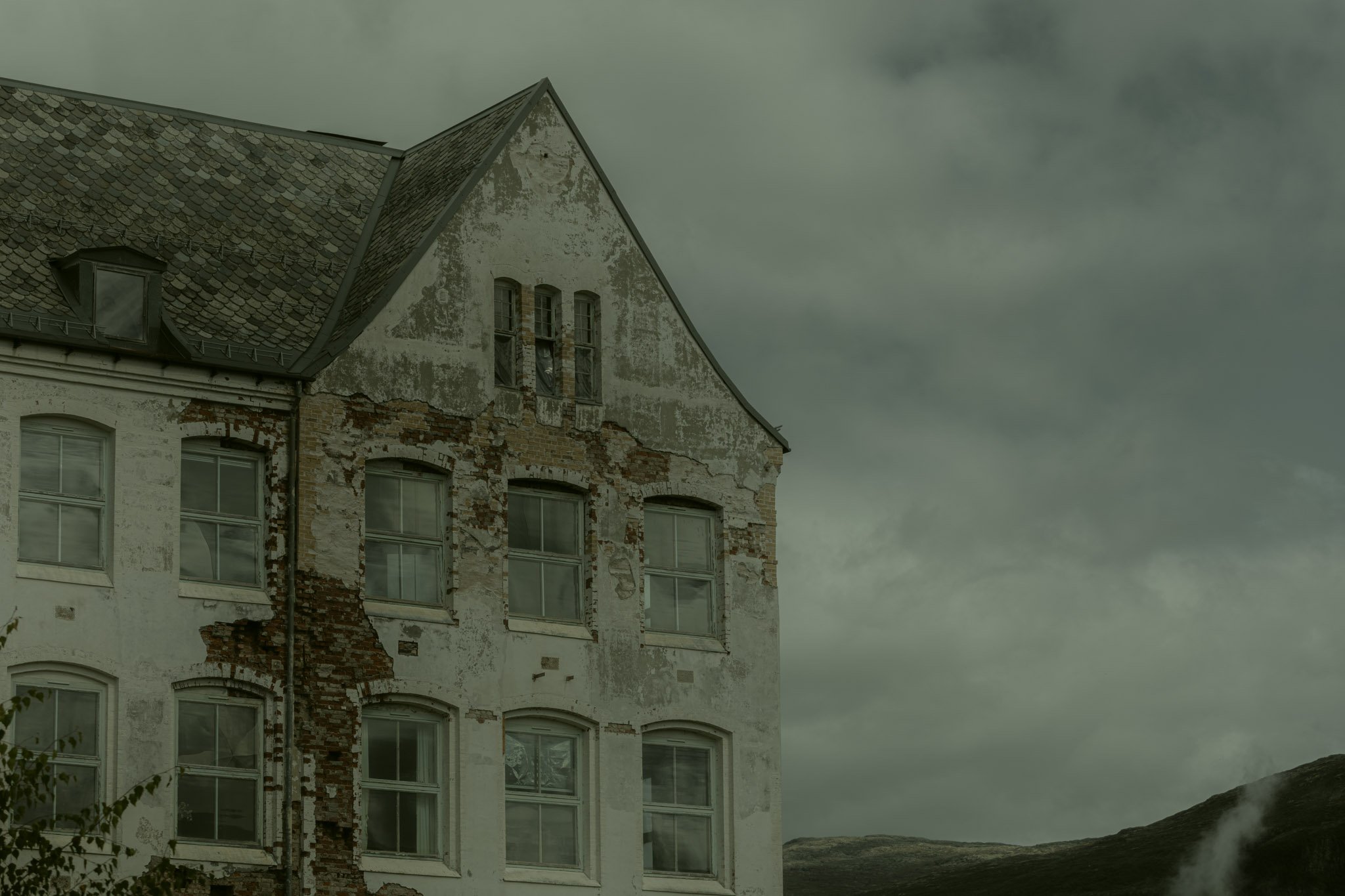 An old, weathered building with peeling paint, exposed bricks, and multiple windows under a dark, cloudy sky.