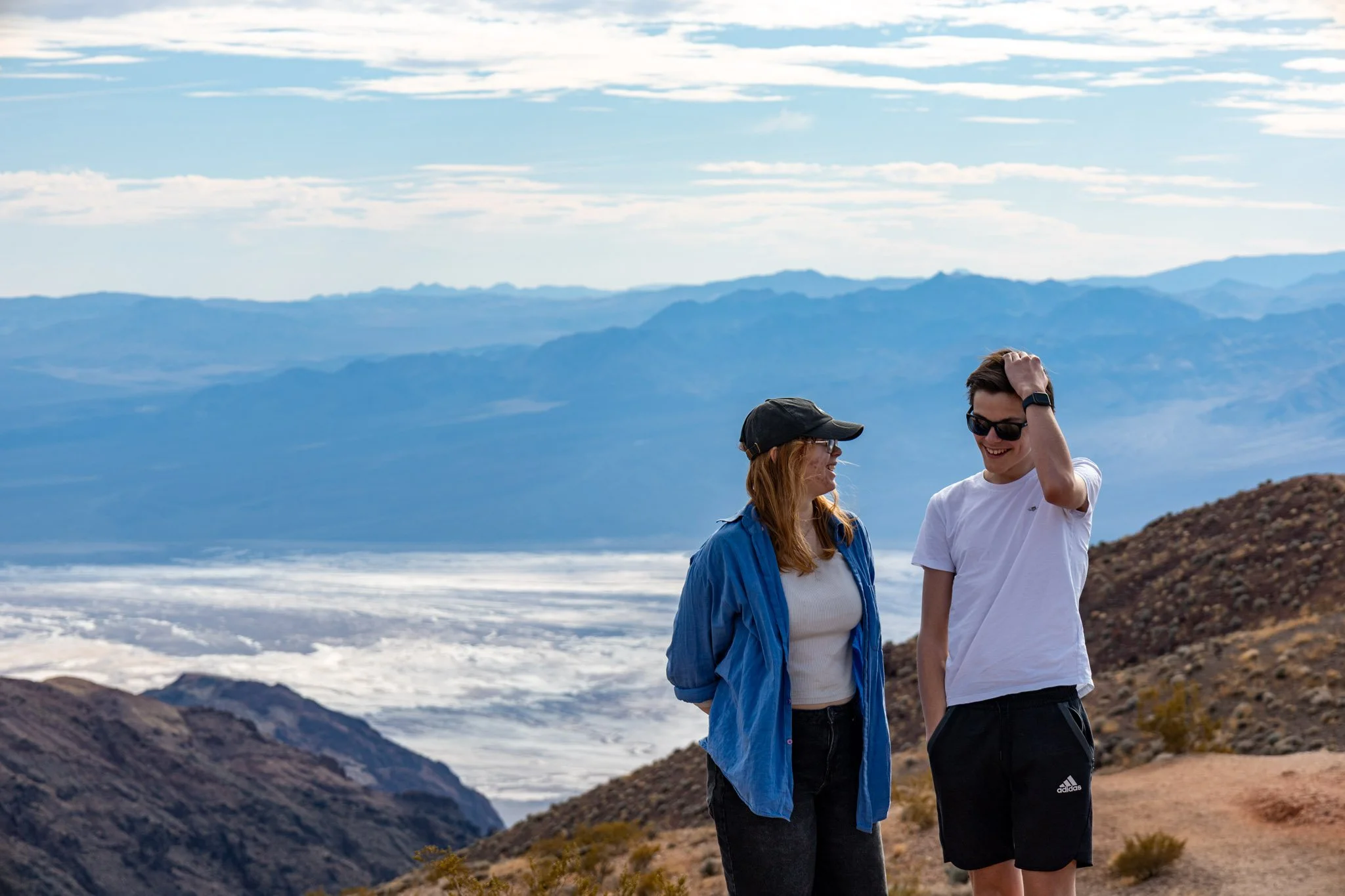 A man and woman standing outdoors on a mountain trail, smiling with mountains and a cloudy sky in the background.