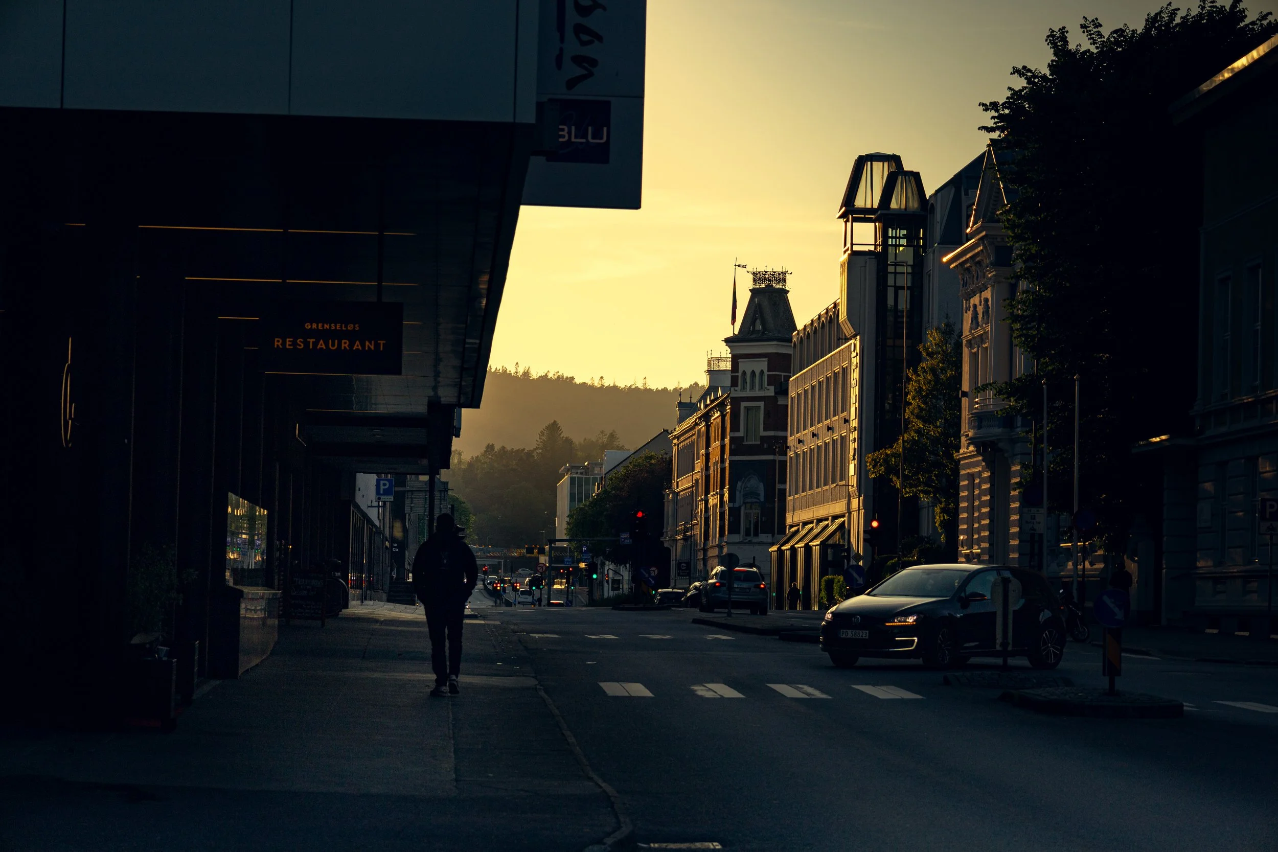 City street at sunset with cars parked along the road, a pedestrian walking, and historic-style buildings with turrets and decorative details on the right side.