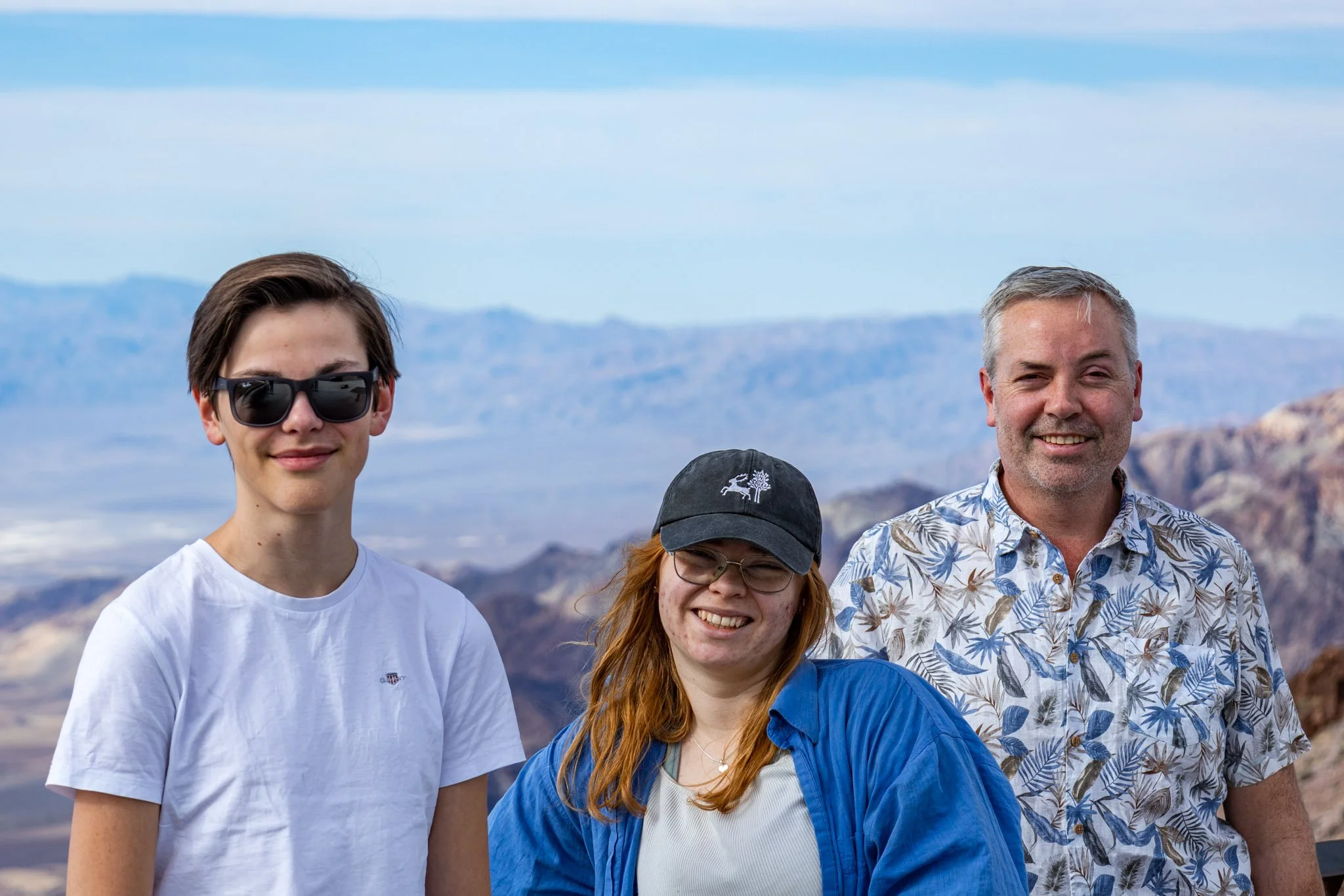 Group of four people smiling outdoors with a mountainous landscape in the background. The group includes a young man with dark sunglasses and a white t-shirt, a red-haired woman with glasses and a blue jacket, an older man with gray hair and a floral shirt, and part of another person wearing a dark cap.