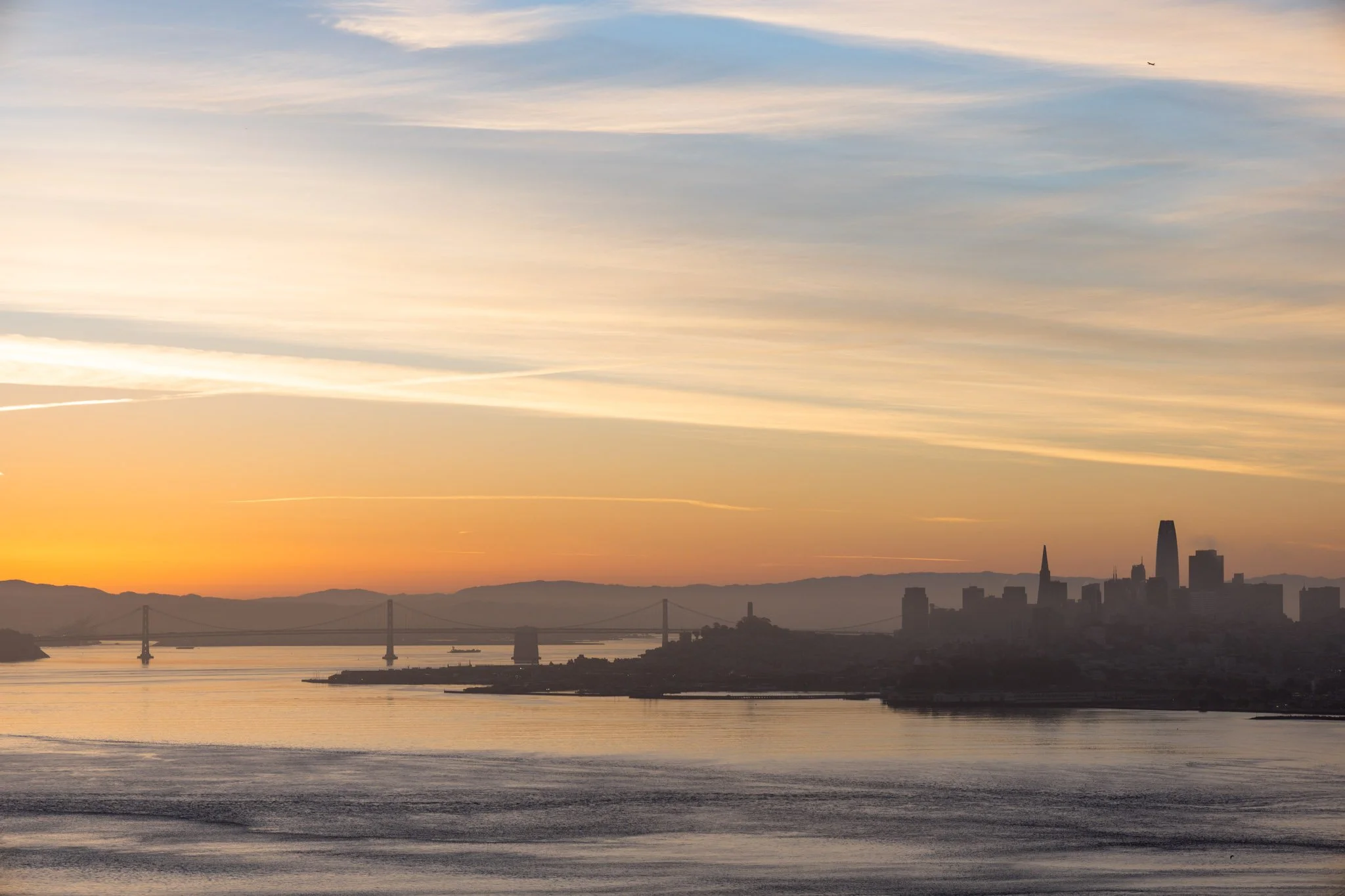A city skyline during sunset with a bridge and water in the foreground, and a sky with light clouds and a small airplane high above.