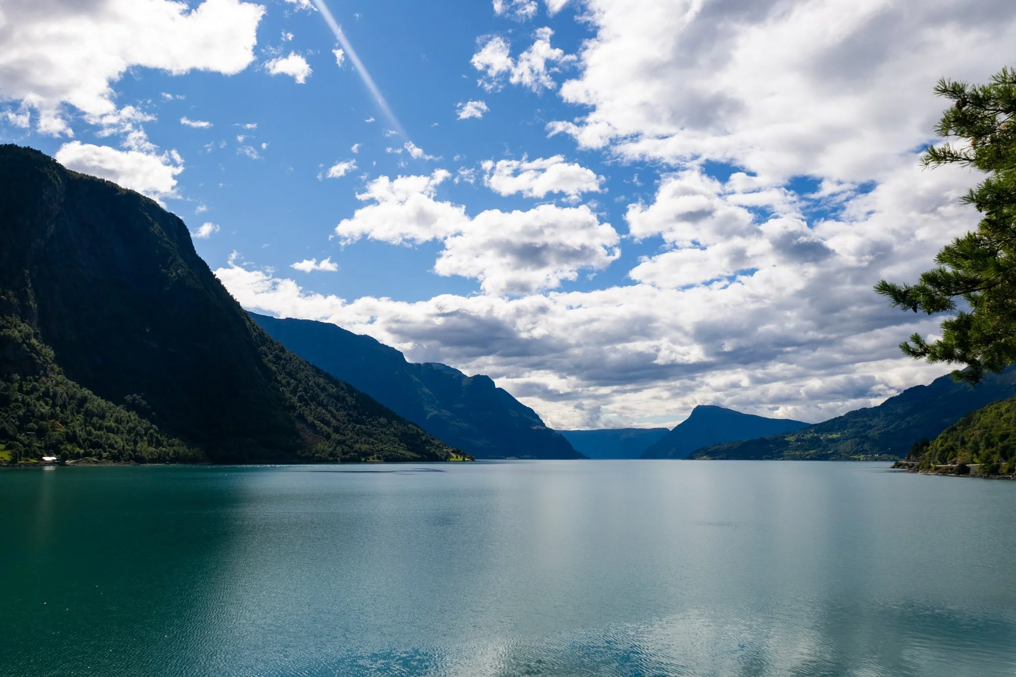 Scenic landscape of a large body of water surrounded by mountains and trees under a partly cloudy sky.