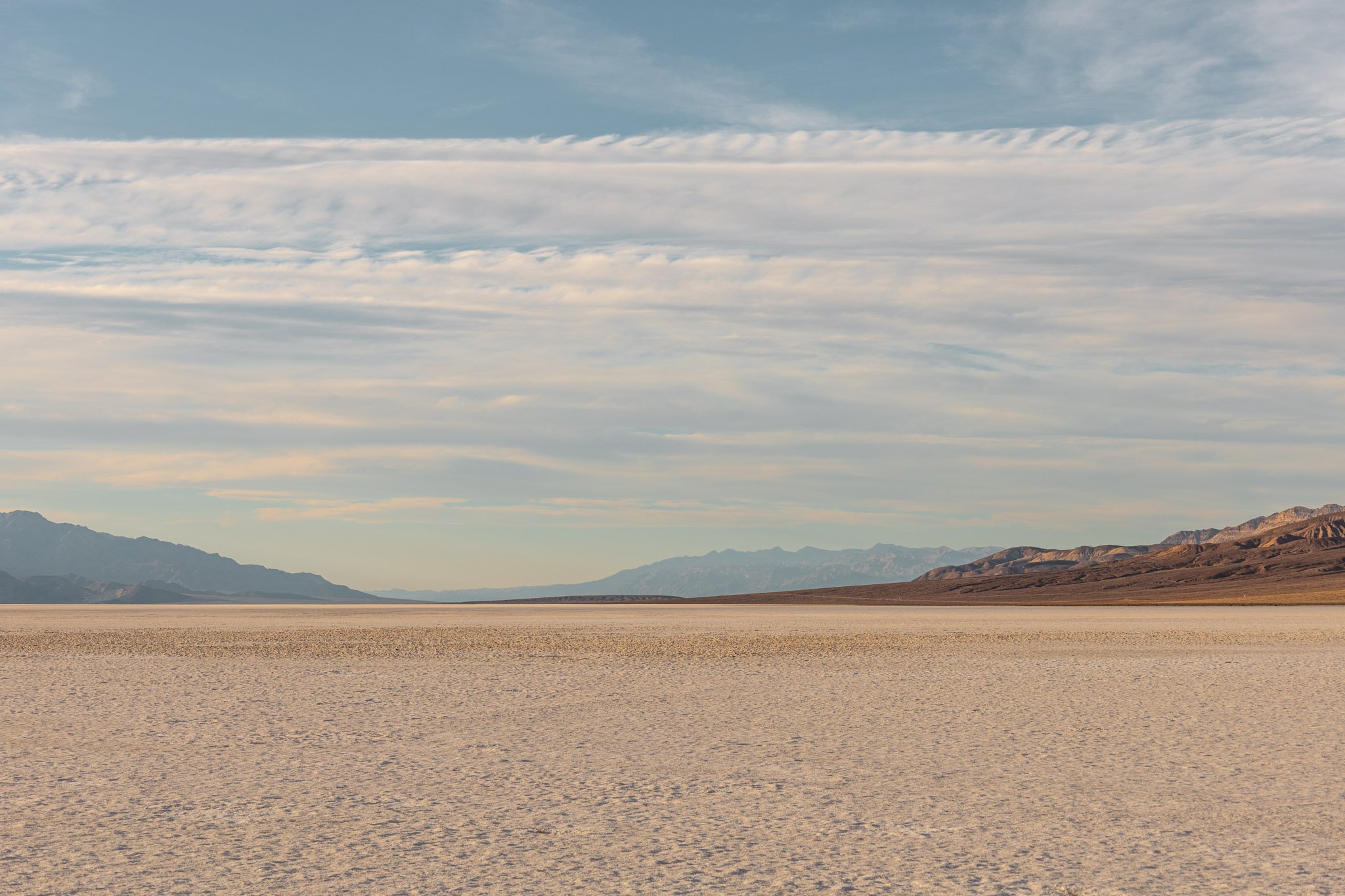 A vast desert landscape with mountains in the distance and a wide, flat expanse in the foreground. The sky above is partly cloudy with light, wispy clouds.