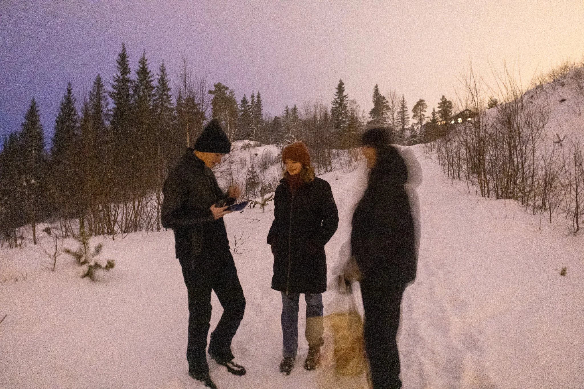 Three people standing in snow outdoors during winter, wearing warm clothing and hats, with a forested hillside and a house with lit windows in the background.