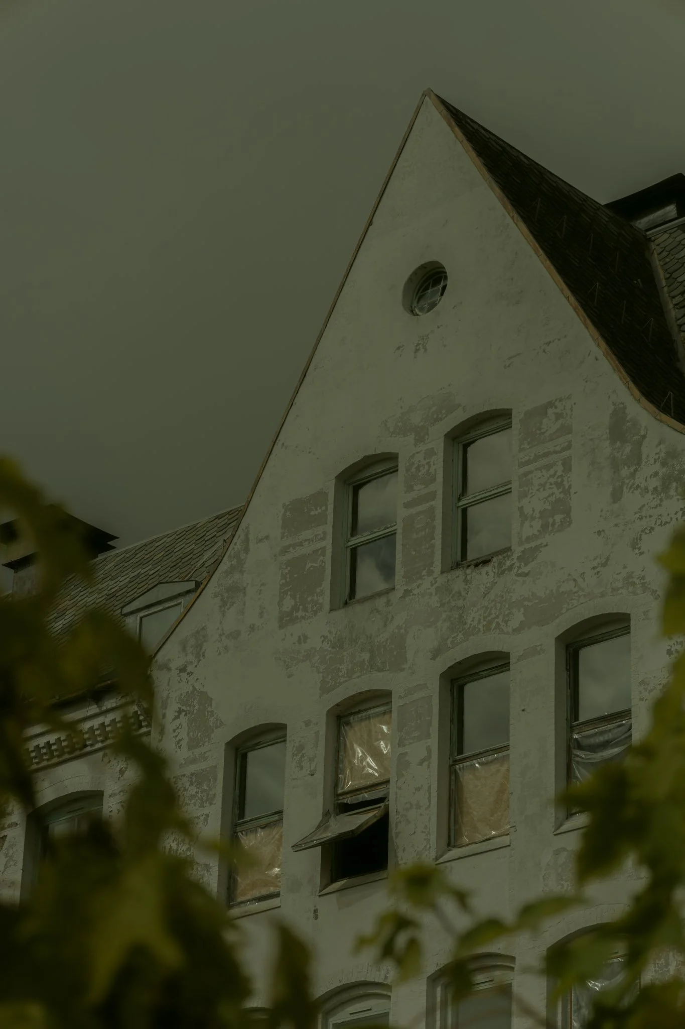 A tall, old building with a steep, dark roof and multiple windows, some of which have plastic coverings. The building has a weathered, white exterior with patches of peeling paint.