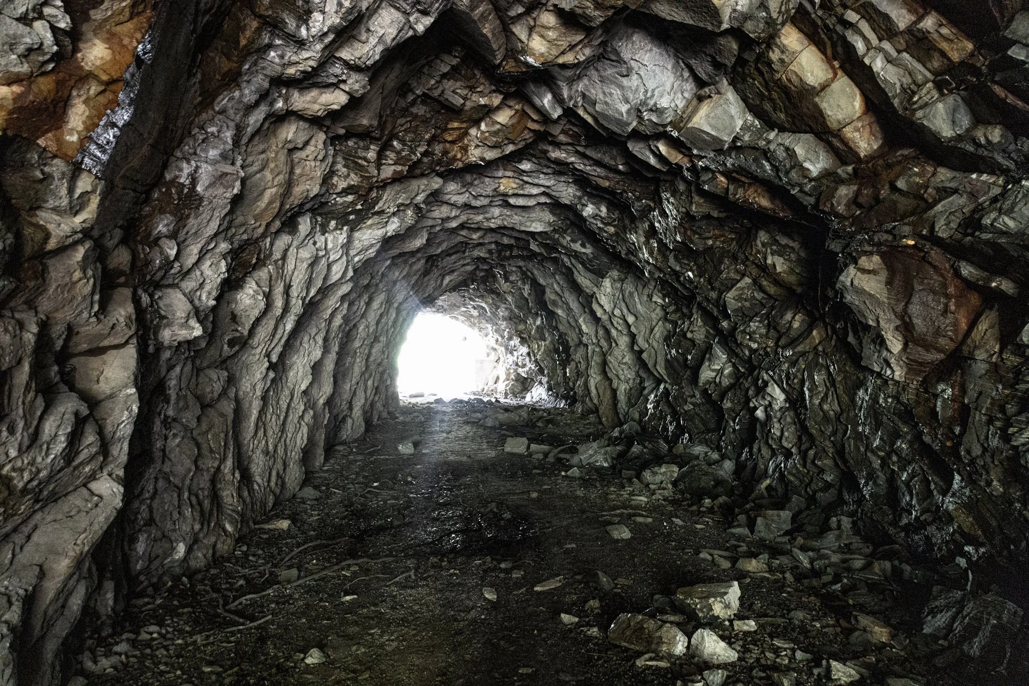 A view inside a rocky tunnel with rugged, uneven walls and ceiling made of dark, layered stones. Light is visible at the tunnel's exit, illuminating part of the dirt ground inside.
