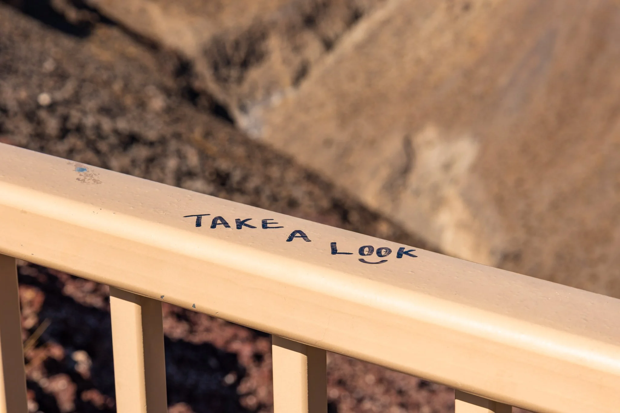 Close-up of a beige wooden railing with the words 'TAKE A LOOK' written in black, with a blurred background of dirt or gravel.