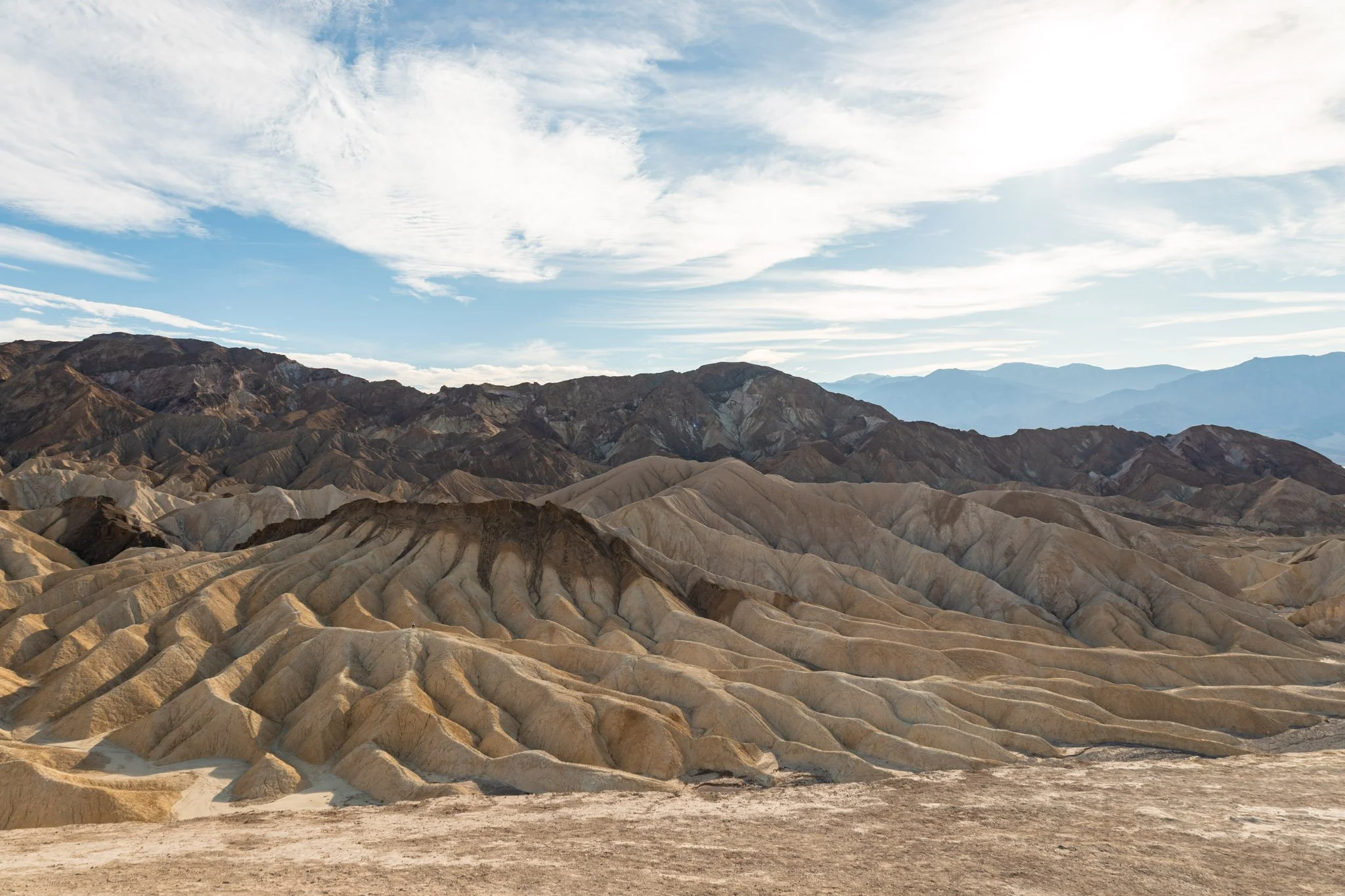 A barren, multicolored desert landscape with rocky, eroded formations under a bright sky with scattered clouds.