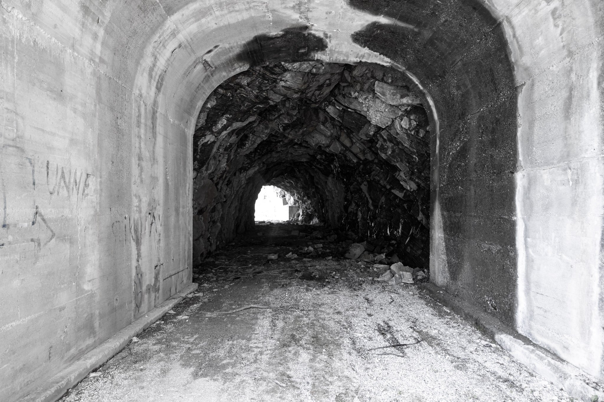 A black and white photograph of a tunnel with a curved ceiling, lined with rocks and concrete, with rubble and debris on the ground.