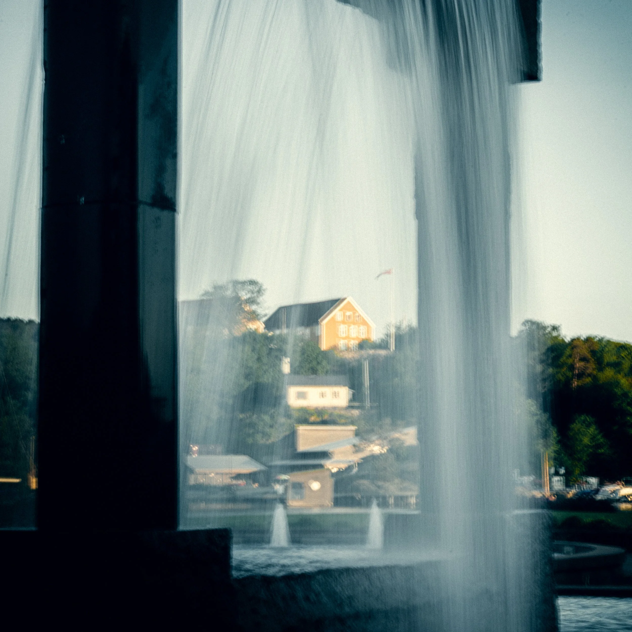 Rainstorm with heavy rainfall over a residential neighborhood, with houses visible in the distance, trees, and cars on the road.
