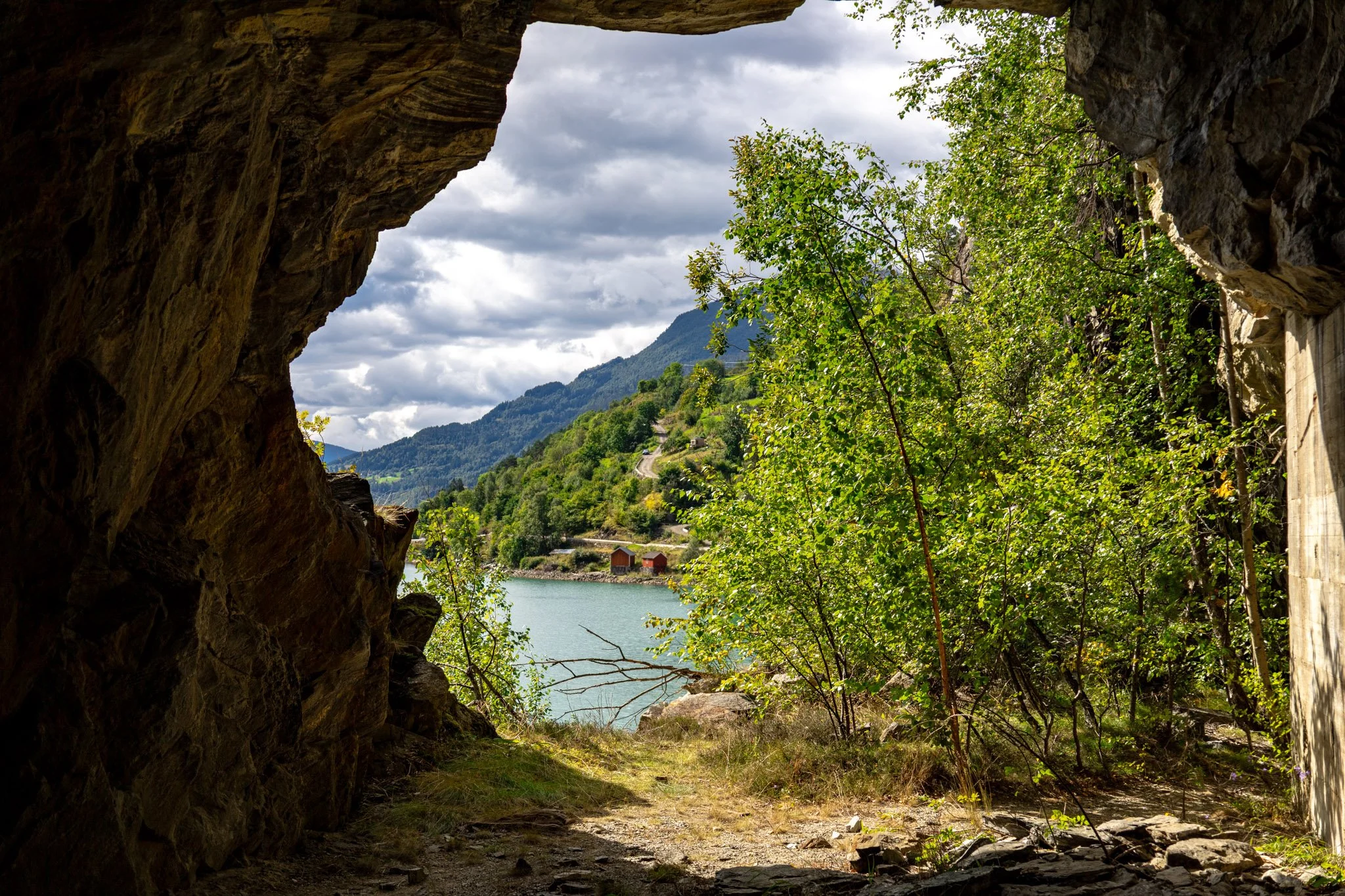 A view from inside a rocky cave looking out at a landscape with lush green trees, mountains in the background, and a lake with small buildings on the shore.