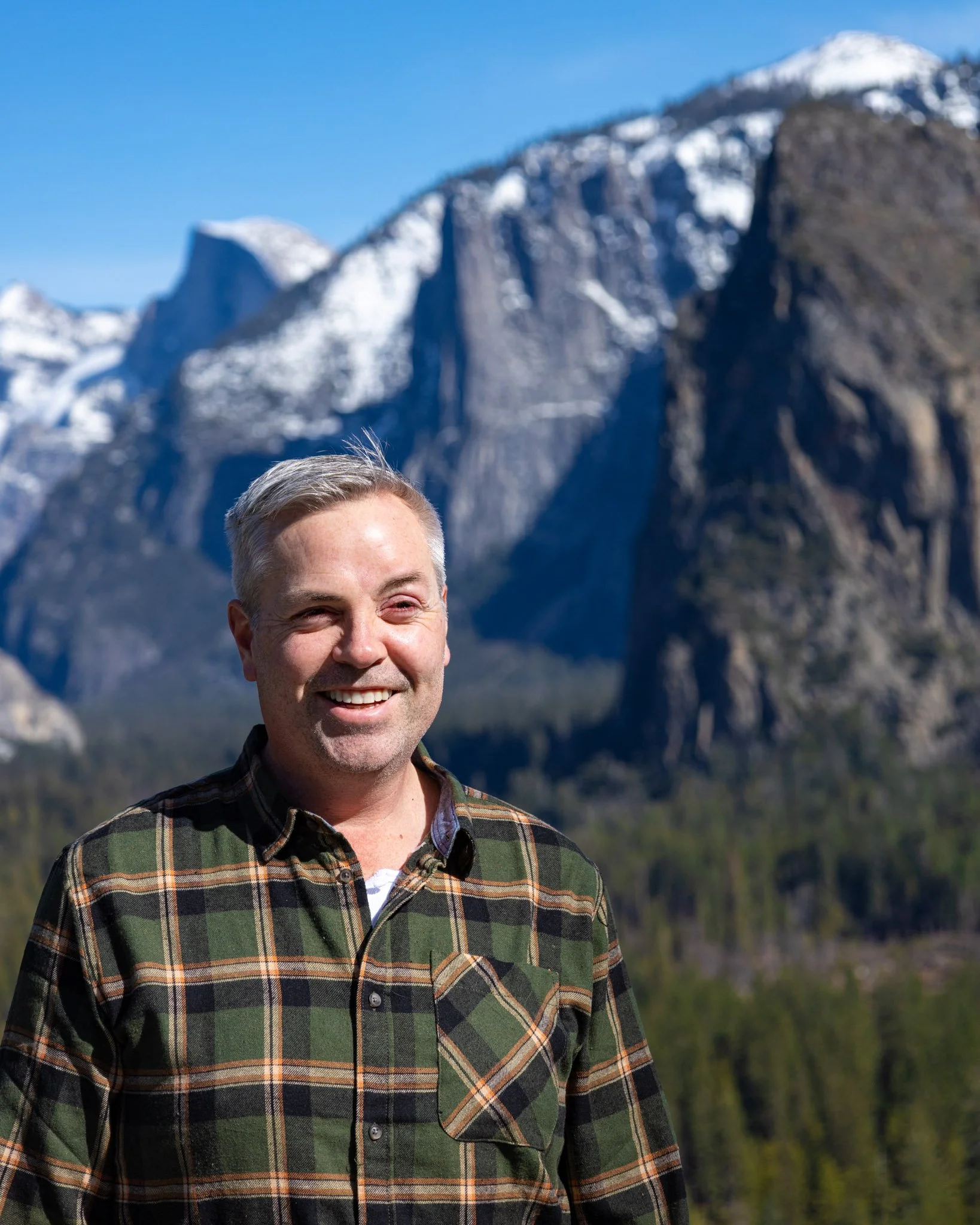 A smiling man with short gray hair wearing a plaid shirt standing outdoors with snow-capped mountains and green trees in the background.