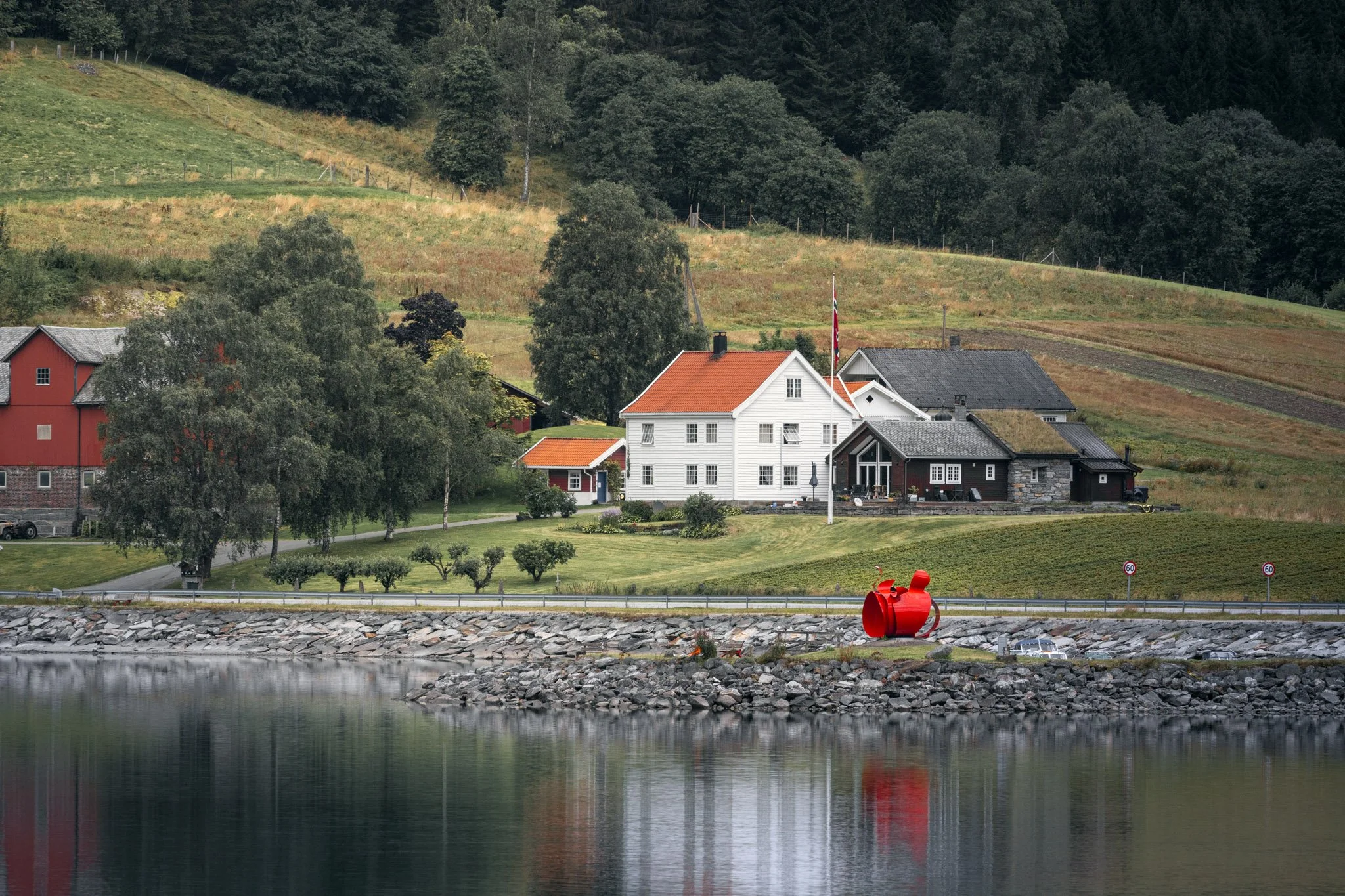 A scenic view of a small seaside village with colorful houses, trees, and a hill in the background, along a rocky shoreline with calm water reflecting the buildings and landscape.