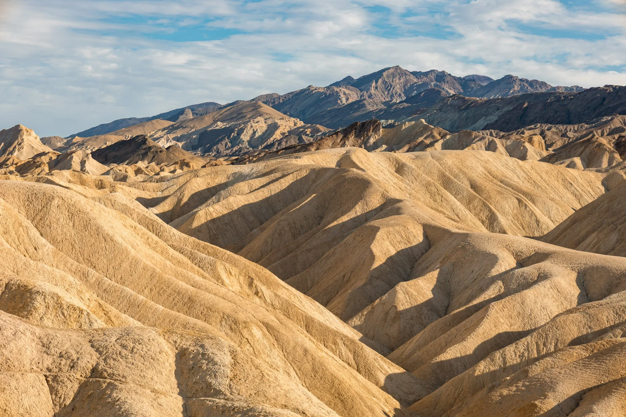 A landscape of rugged, colorful desert mountains with rolling beige and brown hills in the foreground and taller dark and gray mountain peaks in the background under a partly cloudy sky.