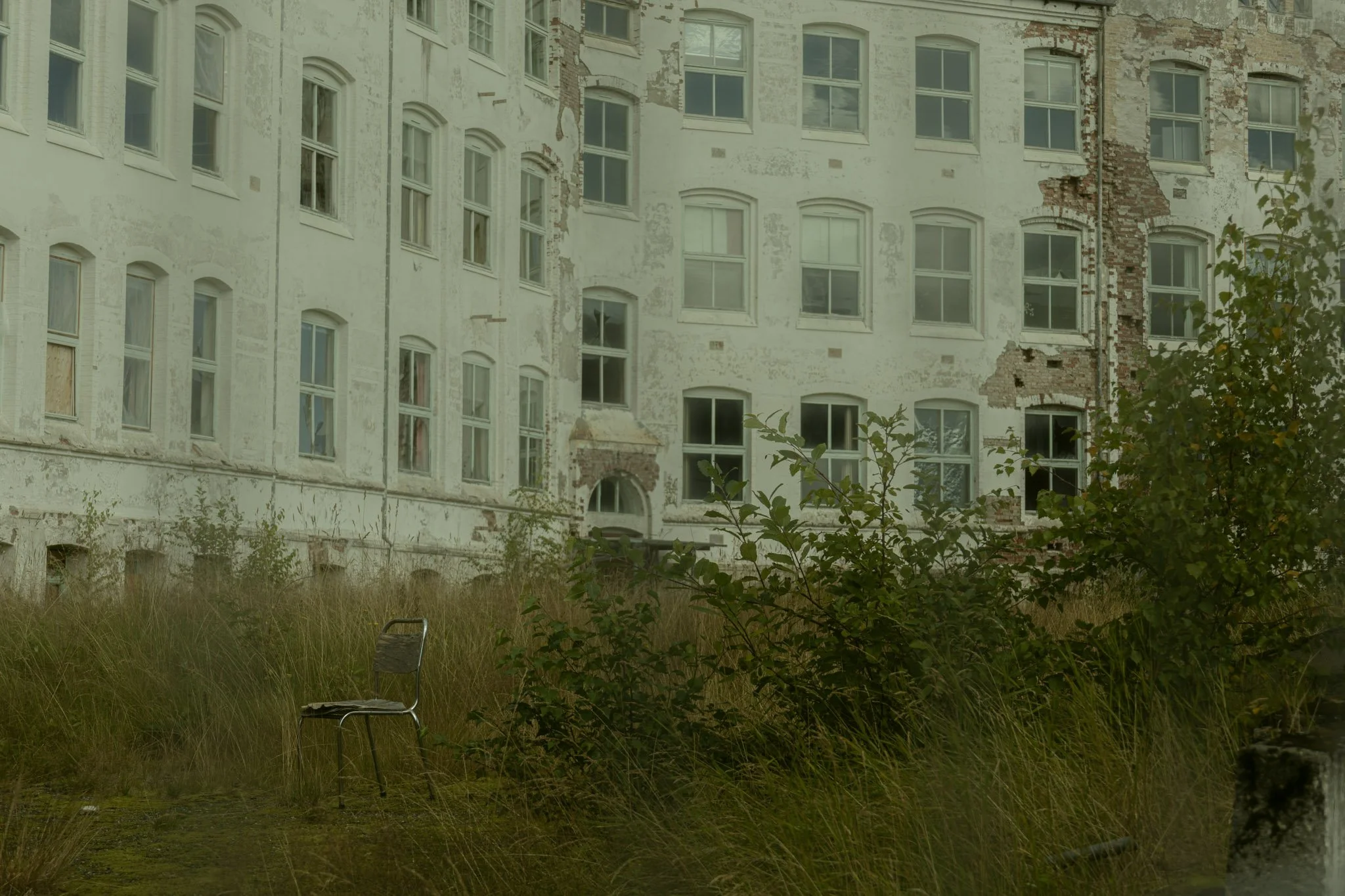 An old, abandoned building with peeling paint and damaged brickwork, surrounded by overgrown grass and bushes, with a metal chair in the foreground.