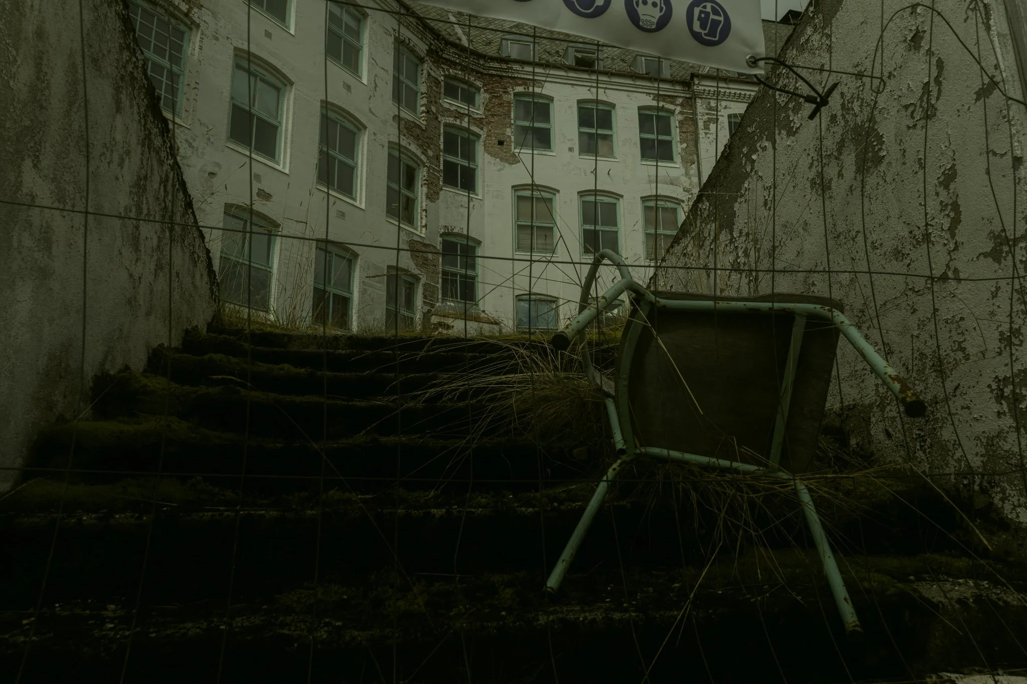A view through a chain-link fence showing an old, weathered building with peeling paint and broken windows, with overgrown grass and a fallen, rusty shopping cart in the foreground.