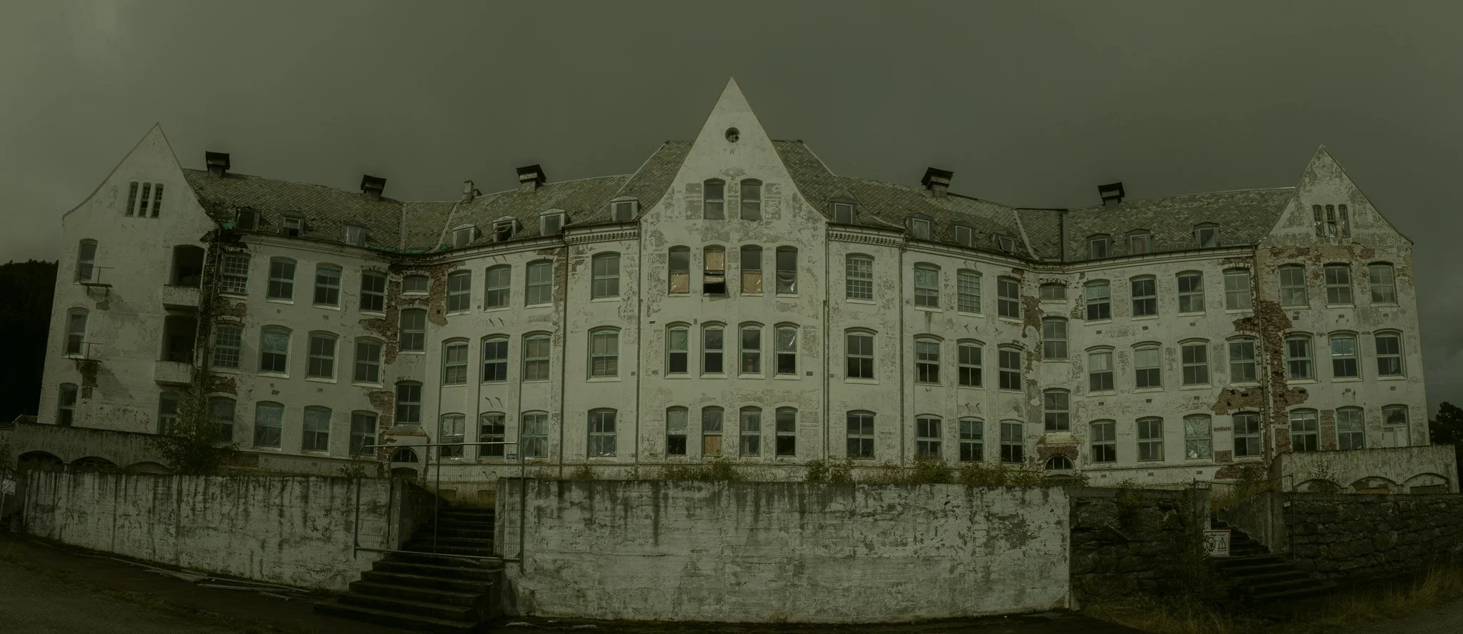 An old, abandoned multi-story building with peeling white paint and broken windows, surrounded by a concrete wall and stairs, under a dark cloudy sky.