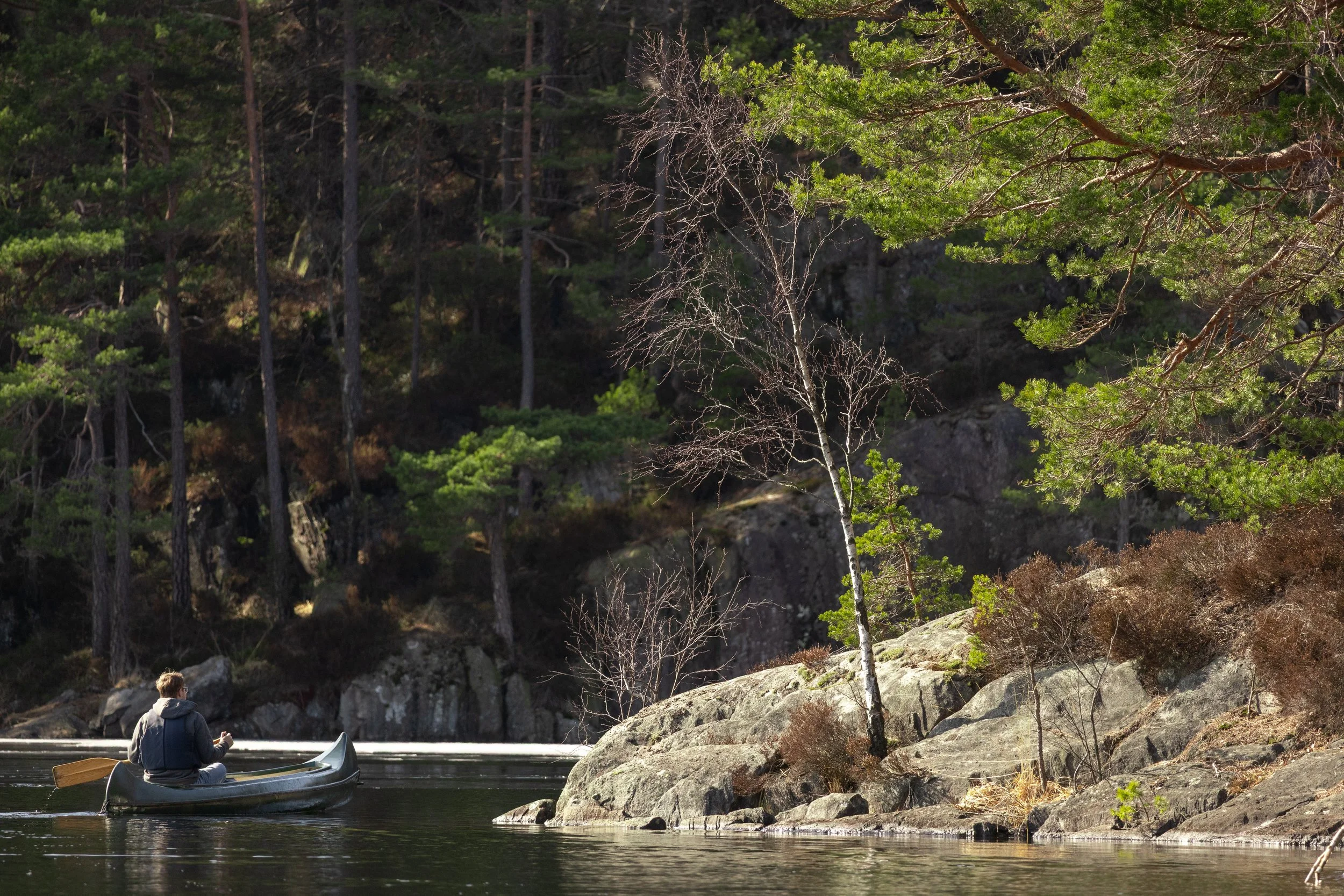 A person sitting in a canoe on a river, surrounded by rocks and trees, with a forested hillside in the background.