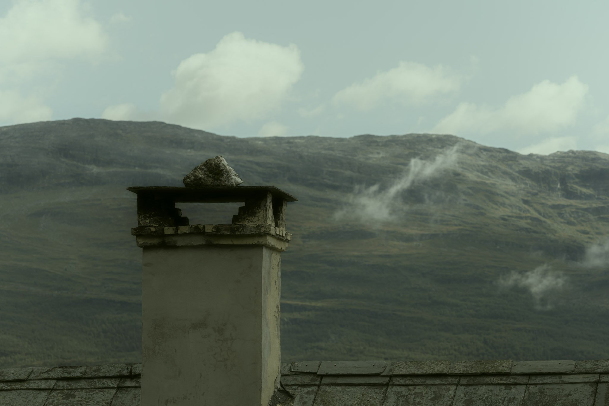 A chimney with smoke rising from it on a house roof, with distant hills and clouds in the background.