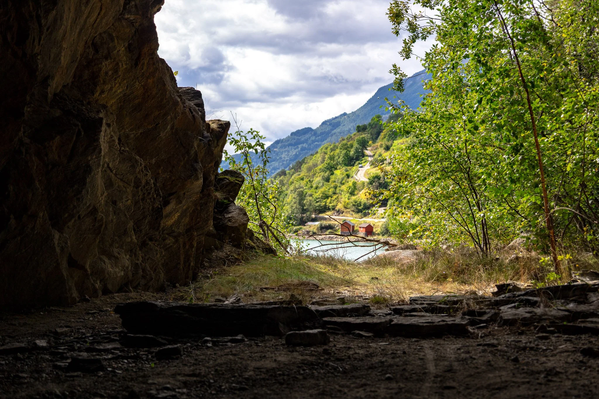 View from inside a cave looking out onto a scenic landscape with green trees, houses, a winding road, a body of water, mountains, and a cloudy sky