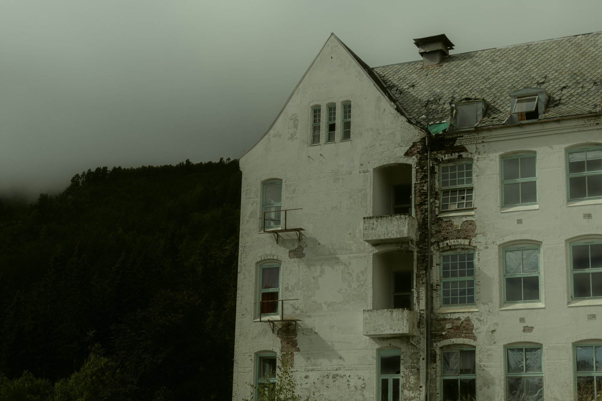 An old, weathered white building with multiple windows, some with broken or missing glass, and visible damage to the exterior wall revealing brickwork underneath. The building is next to a green, forested hillside under an overcast sky.