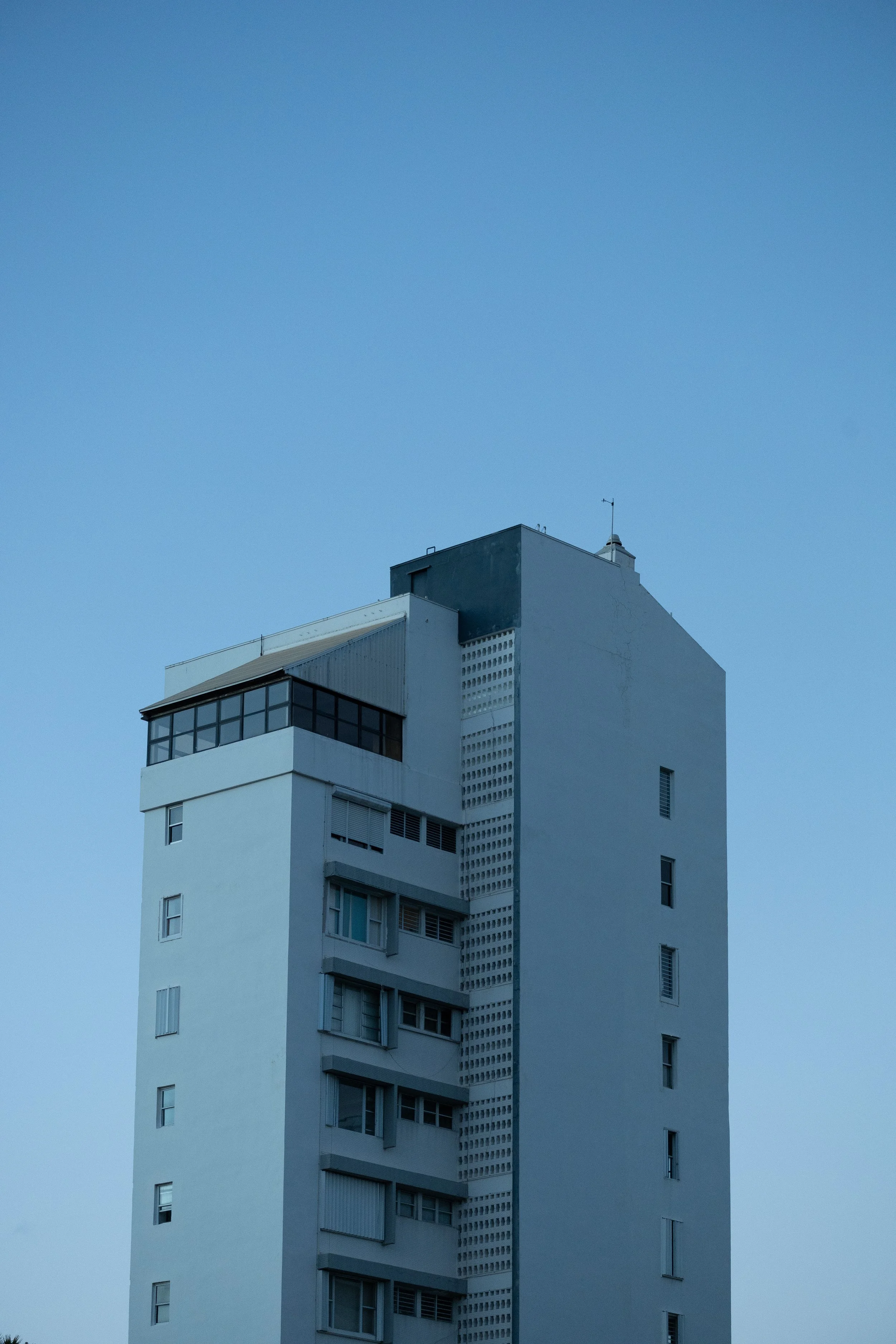A tall white building with multiple balconies and windows, and a smaller black section near the top, against a clear blue sky.