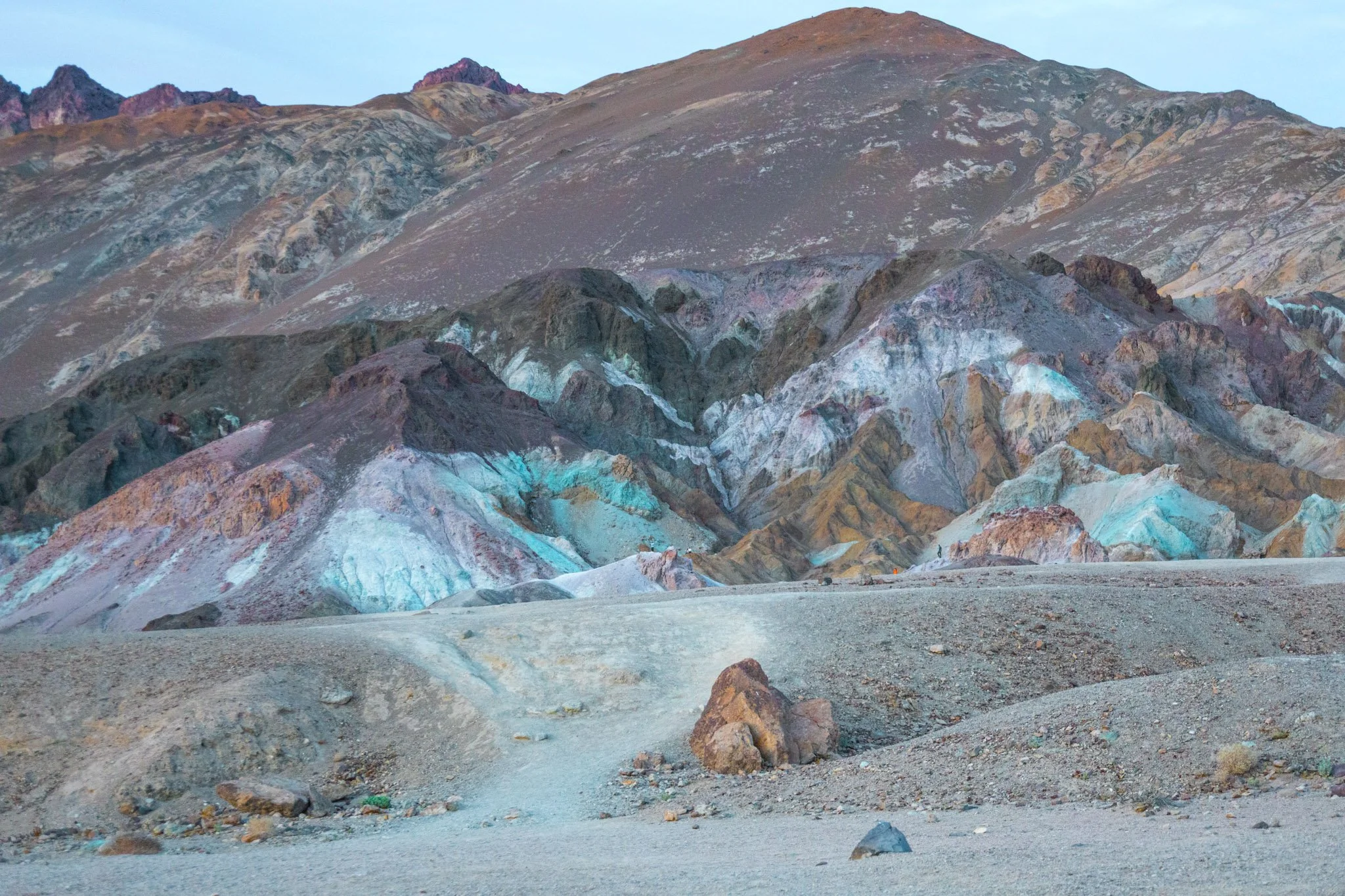 Colorful volcanic rock formations with a barren, dusty landscape in front, and a mountain in the background.