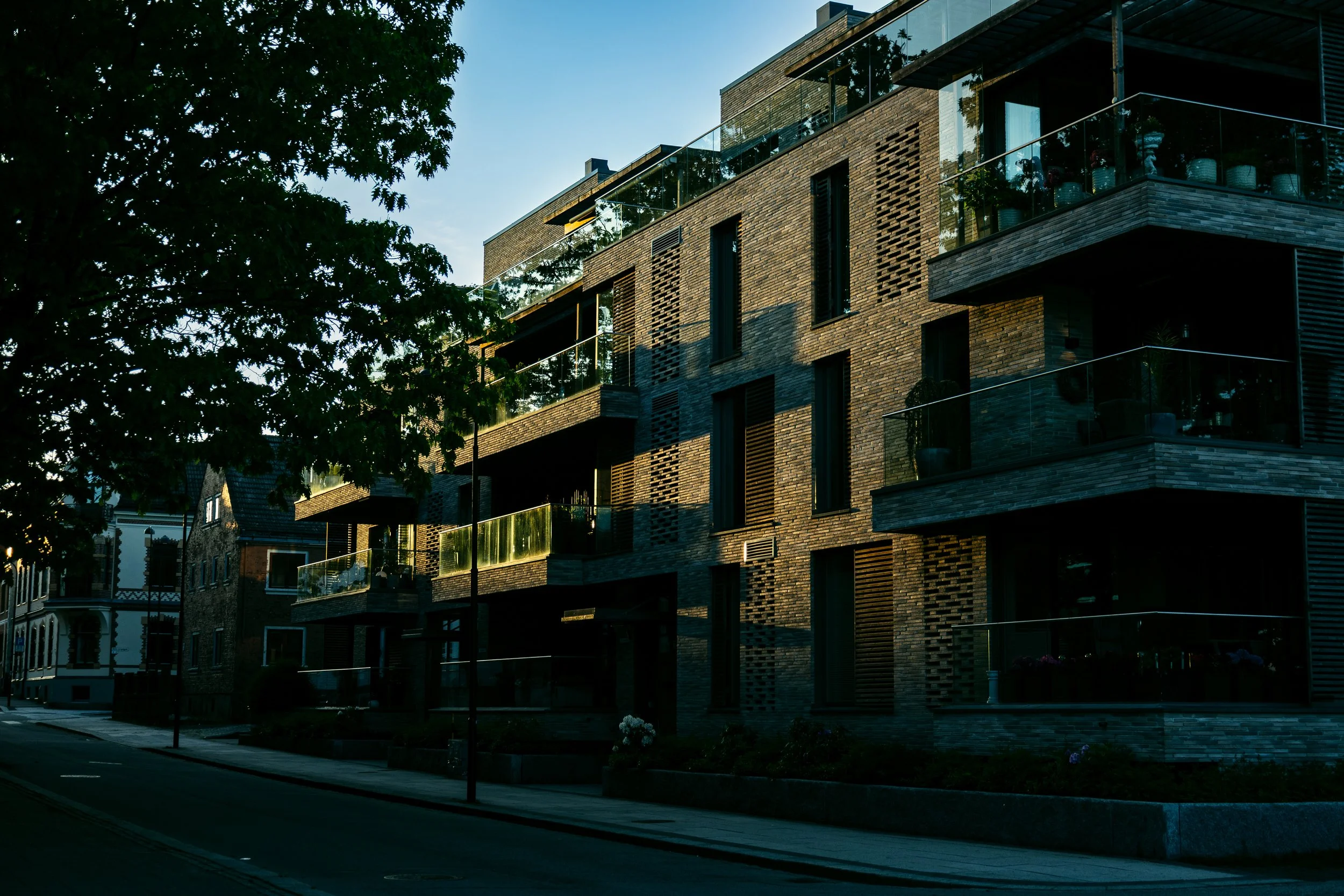 Modern brick building with glass balcony railings and trees in the foreground, during dusk.