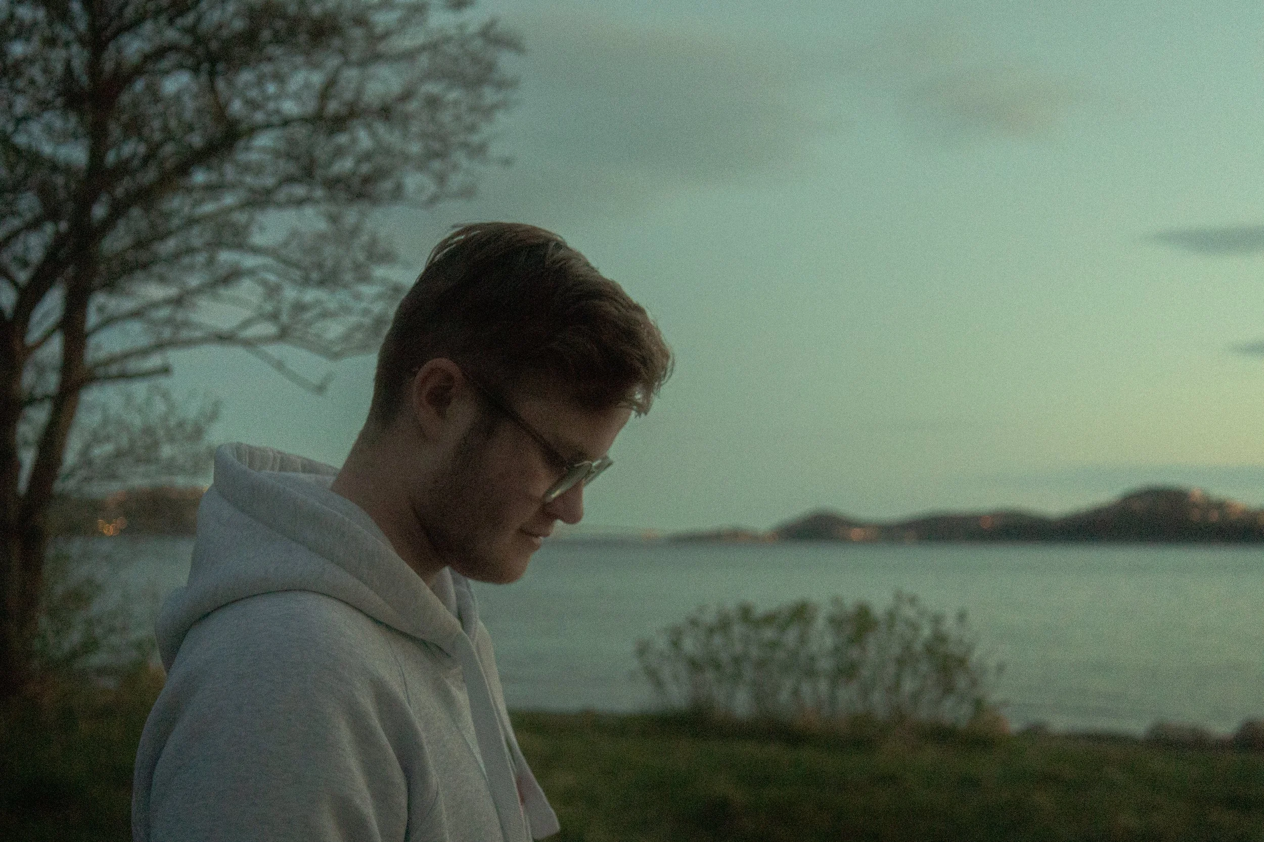 A young man with glasses and reddish hair, wearing a white hoodie, is looking down with a peaceful expression outdoors near a body of water during dusk or dawn. There are trees and hills in the background.