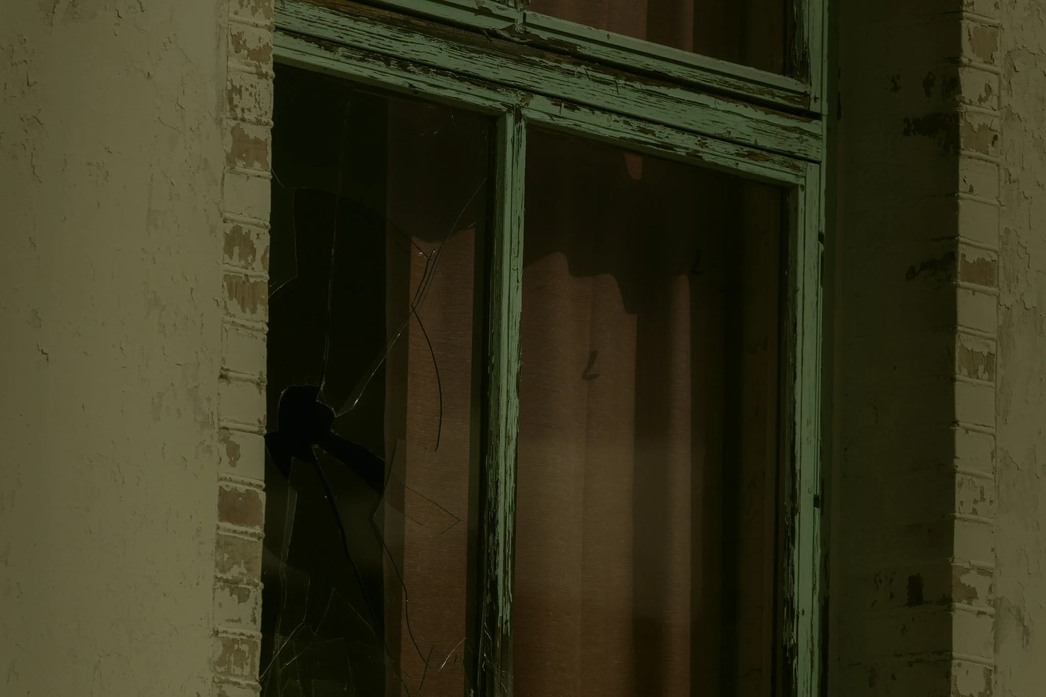 A broken window with a green frame in a peeling yellow wall, showing cracked glass and a dark interior.