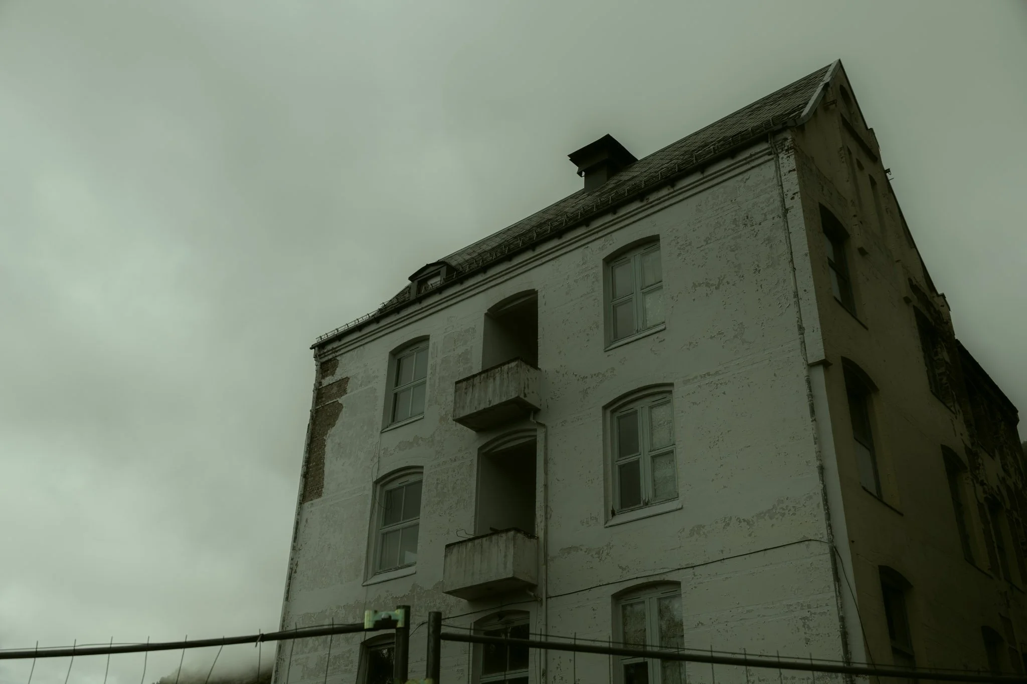 A multi-story old white building with several windows and balconies, showing signs of wear and weathering, under a cloudy sky.