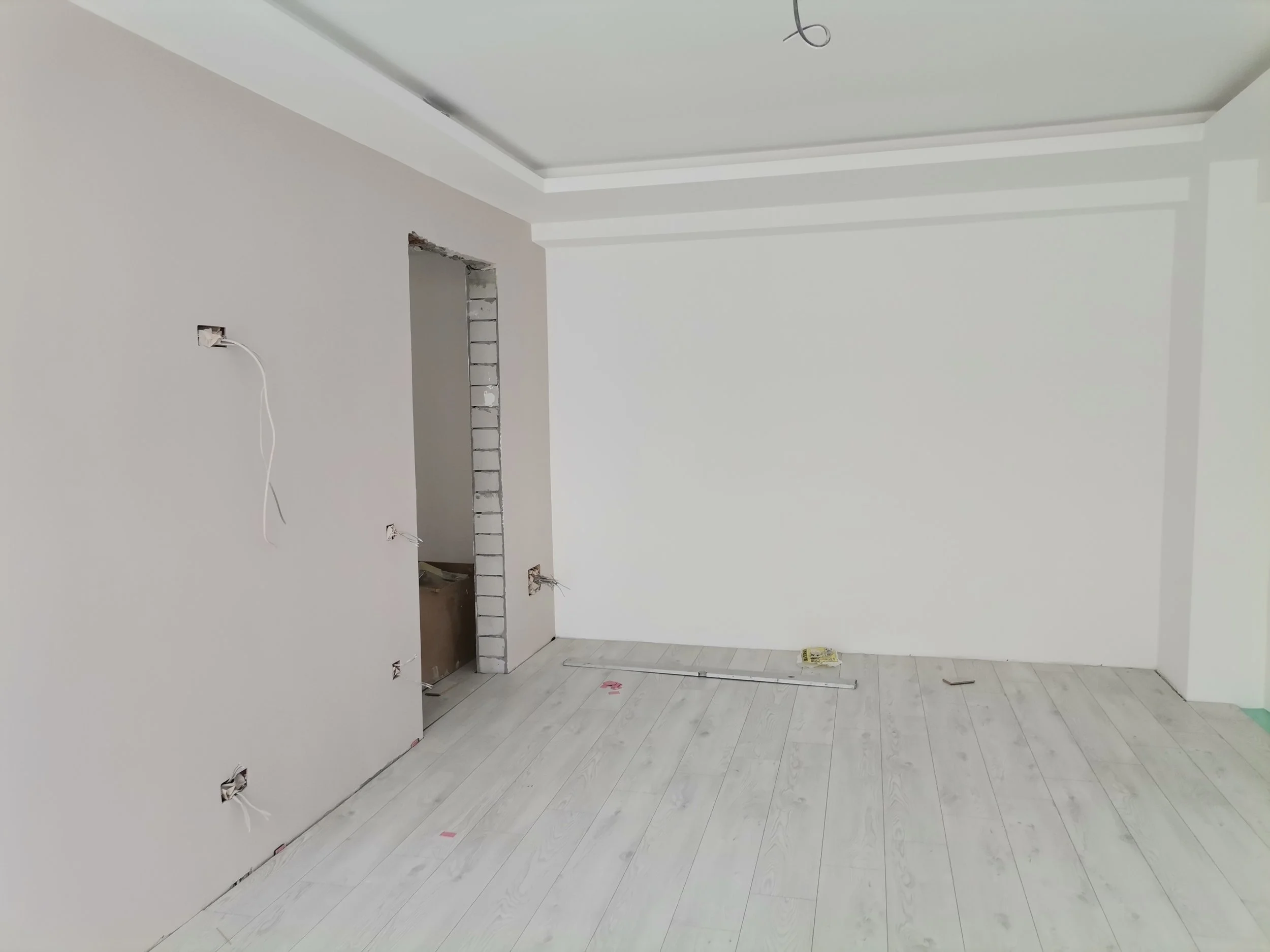 An unfinished room with white walls and flooring, visible electrical wiring, and exposed wall framing. There's construction debris and tools on the floor.