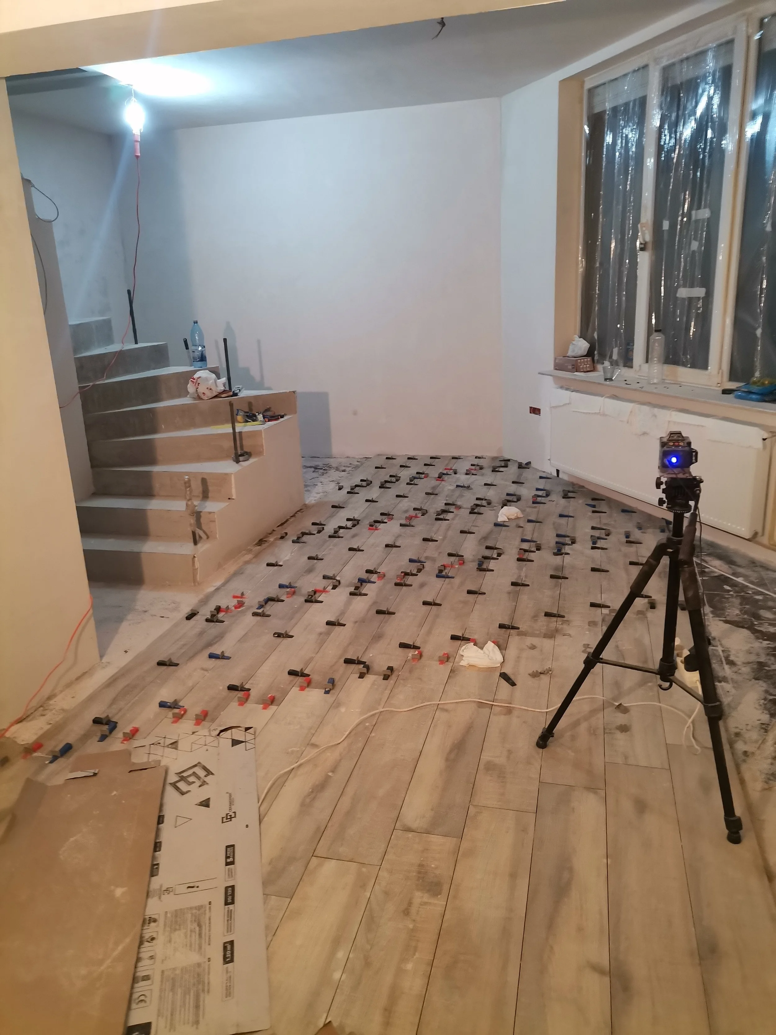 Room under renovation with scattered tools, flooring installation in progress, tripod with camera, and unfinished staircase.