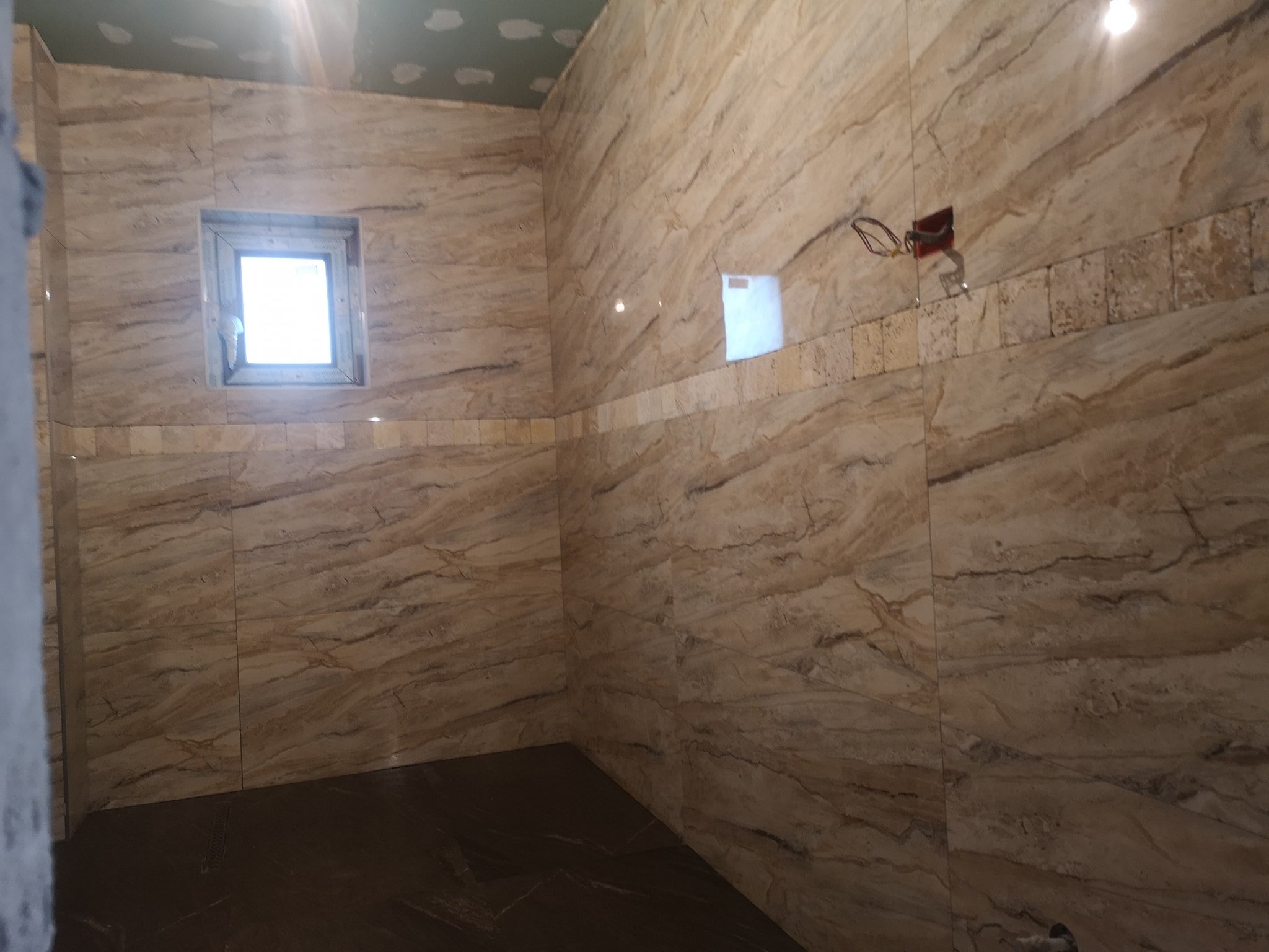 A bathroom with marble-tiled walls and a small window near the ceiling, under construction or renovation with exposed electrical wiring.
