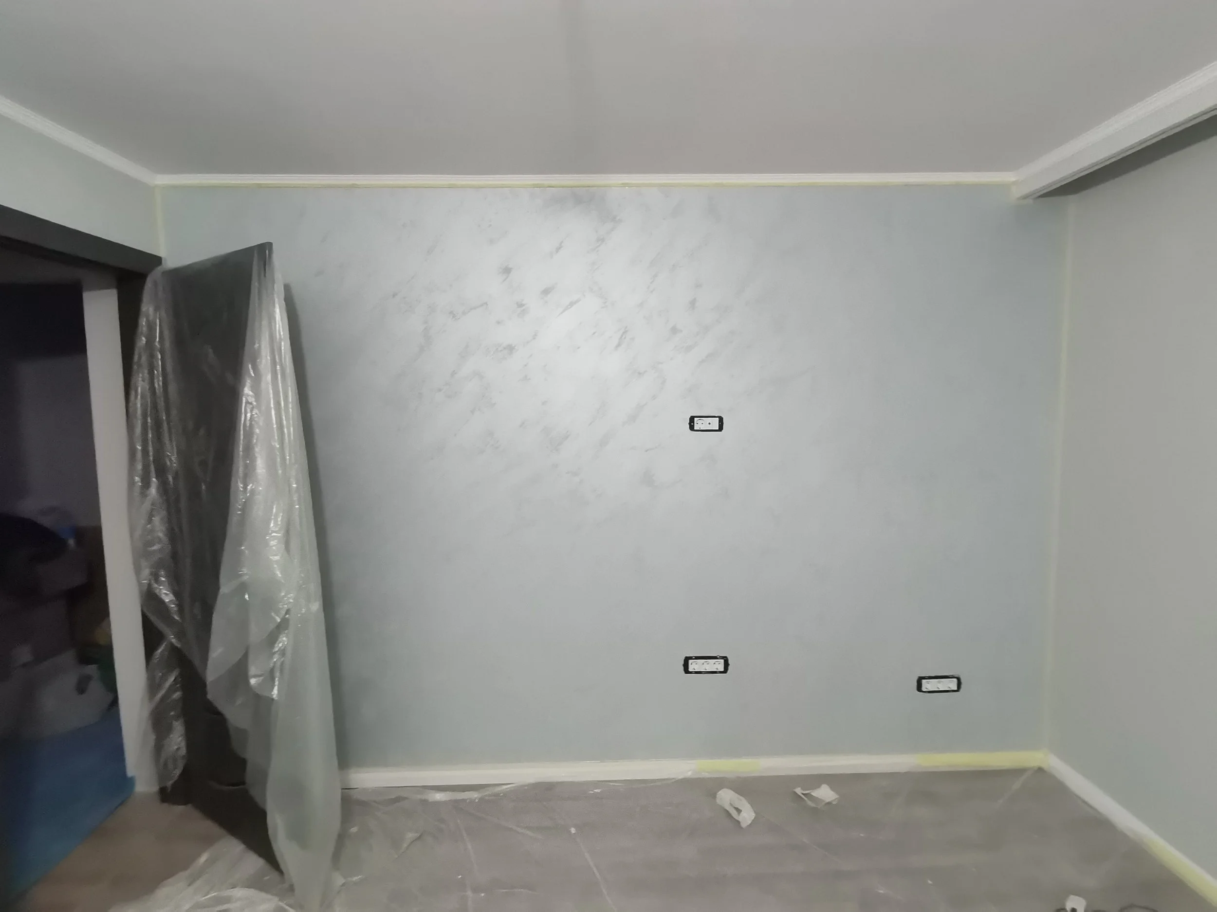 Empty room with painted walls, electrical outlets, covered furniture, and protective plastic covering on the floor and furniture indicating ongoing painting or renovation.