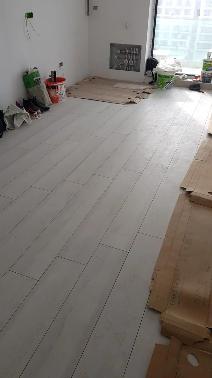 Empty room under renovation with white tiled flooring, construction tools, and supplies along the walls, and a large window with cityscape view.