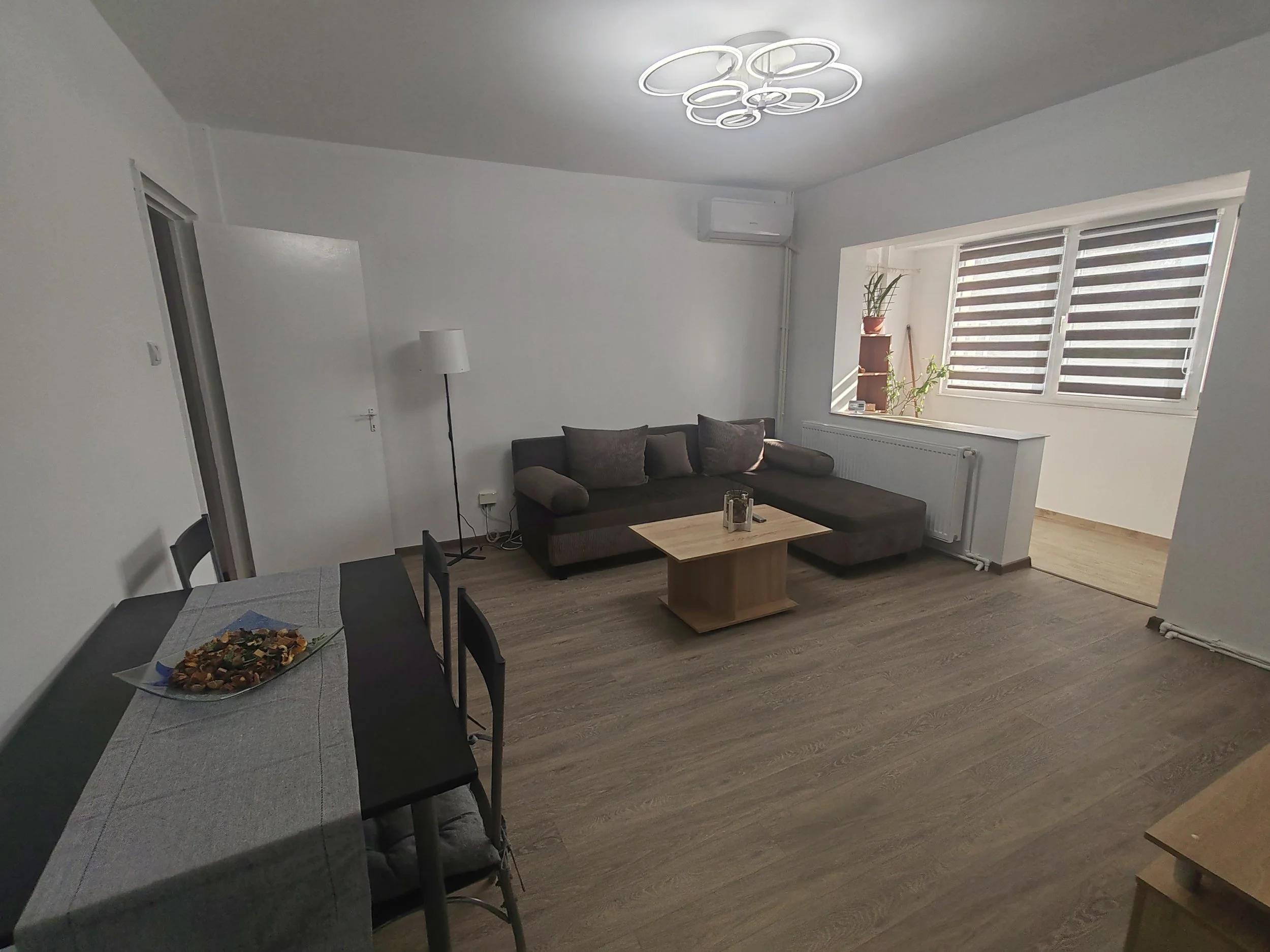 Living room with white walls, large window with blinds, gray sectional sofa with pillows, wooden coffee table, wall-mounted air conditioner, potted plant, dining table with chairs, and a serving tray of snacks.