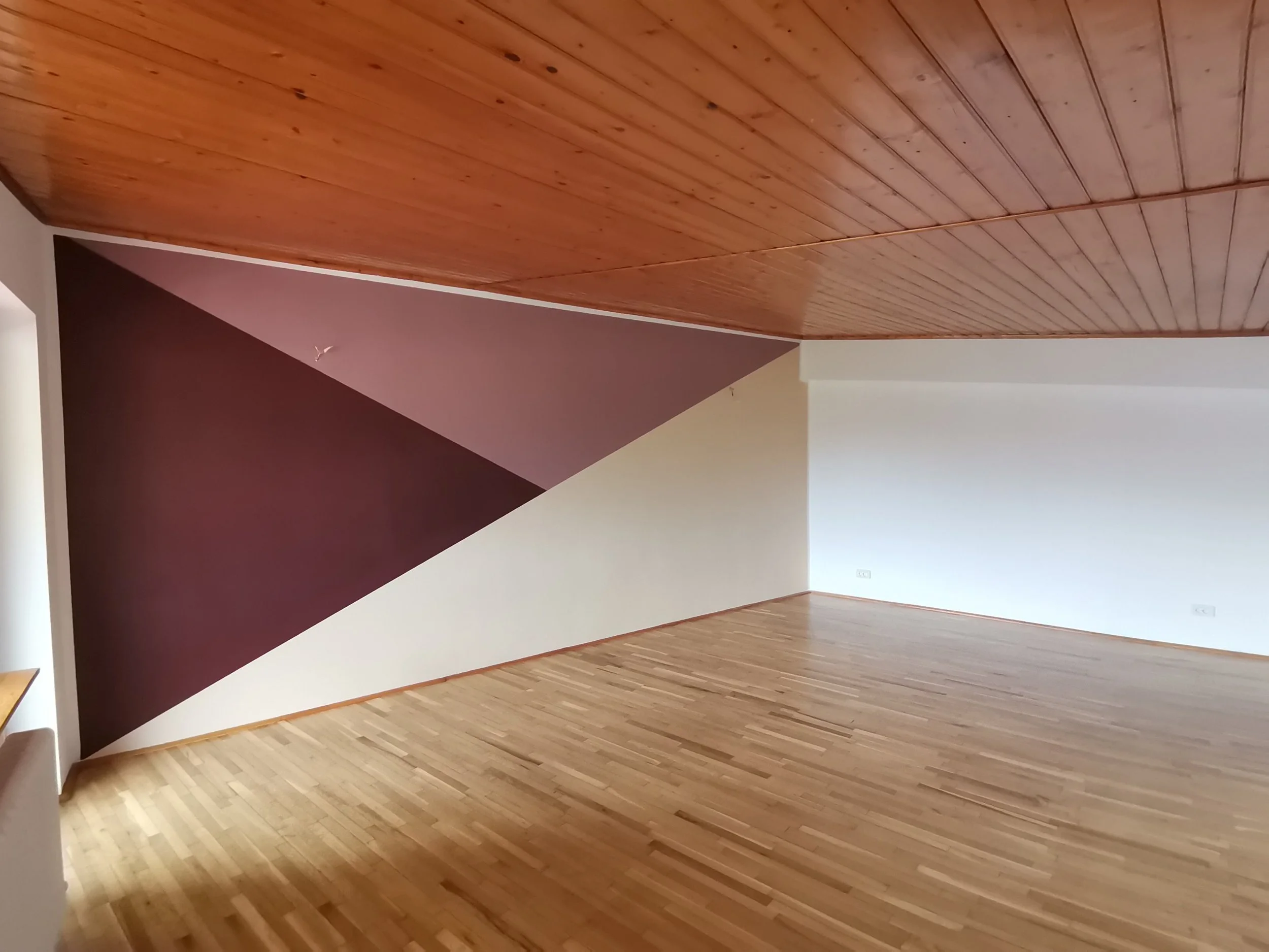 Interior of a room with a wooden ceiling, wood flooring, and walls painted in brown, white, and a pale color. There's a small white electrical outlet on the wall near the floor.